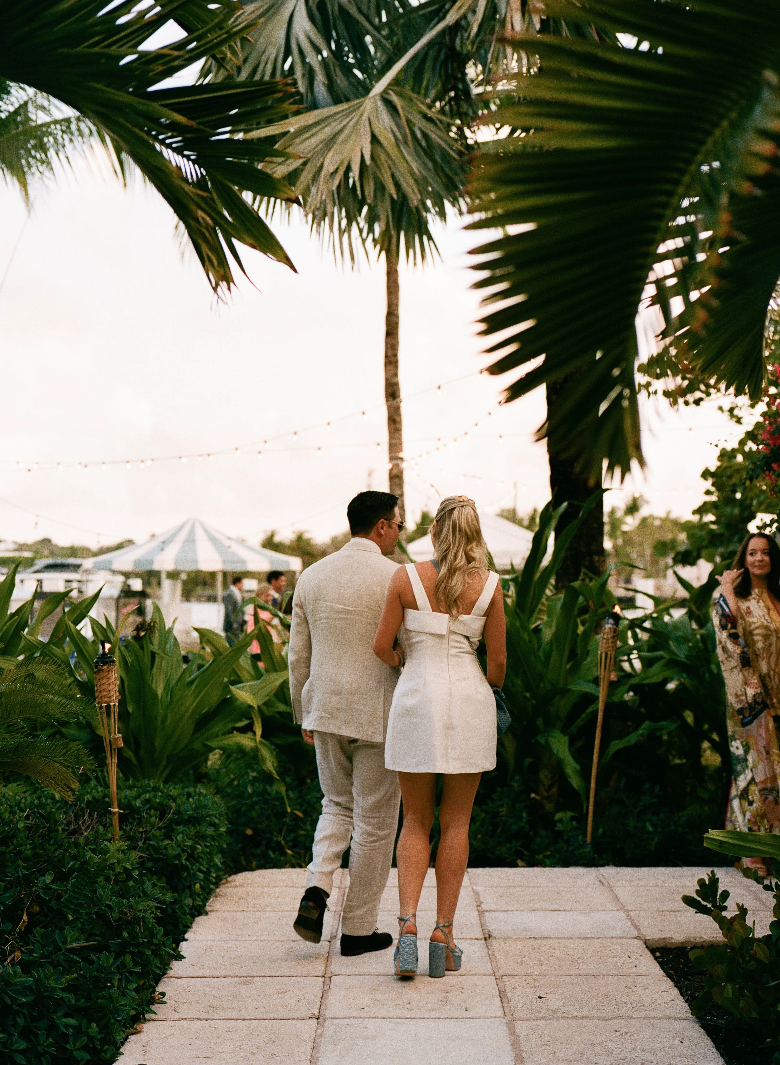 A couple walking on a pathway surrounded by lush tropical greenery at an outdoor event during sunset, with string lights overhead and a woman standing to the right.
