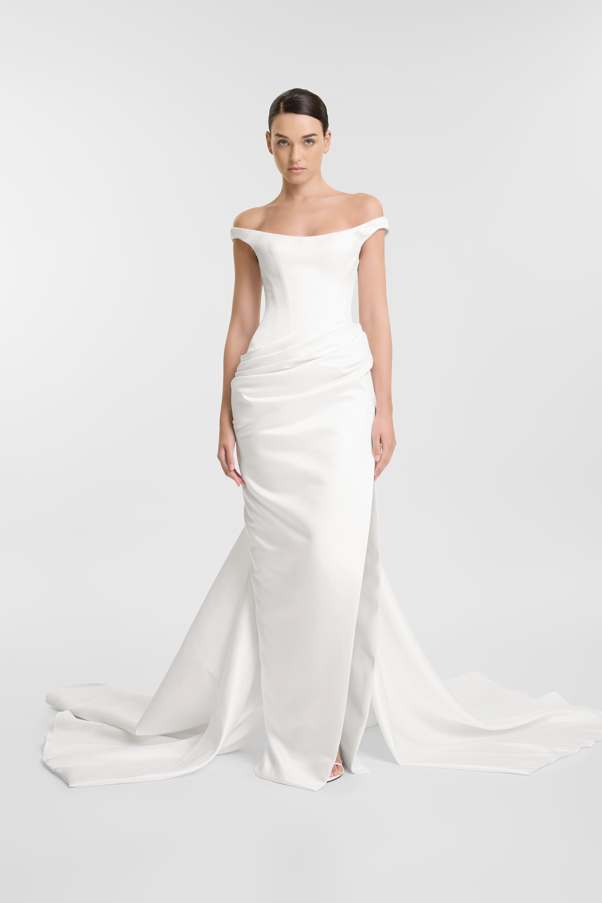 A woman wearing a white off-the-shoulder wedding dress with a fitted bodice and long train, standing against a plain white background.