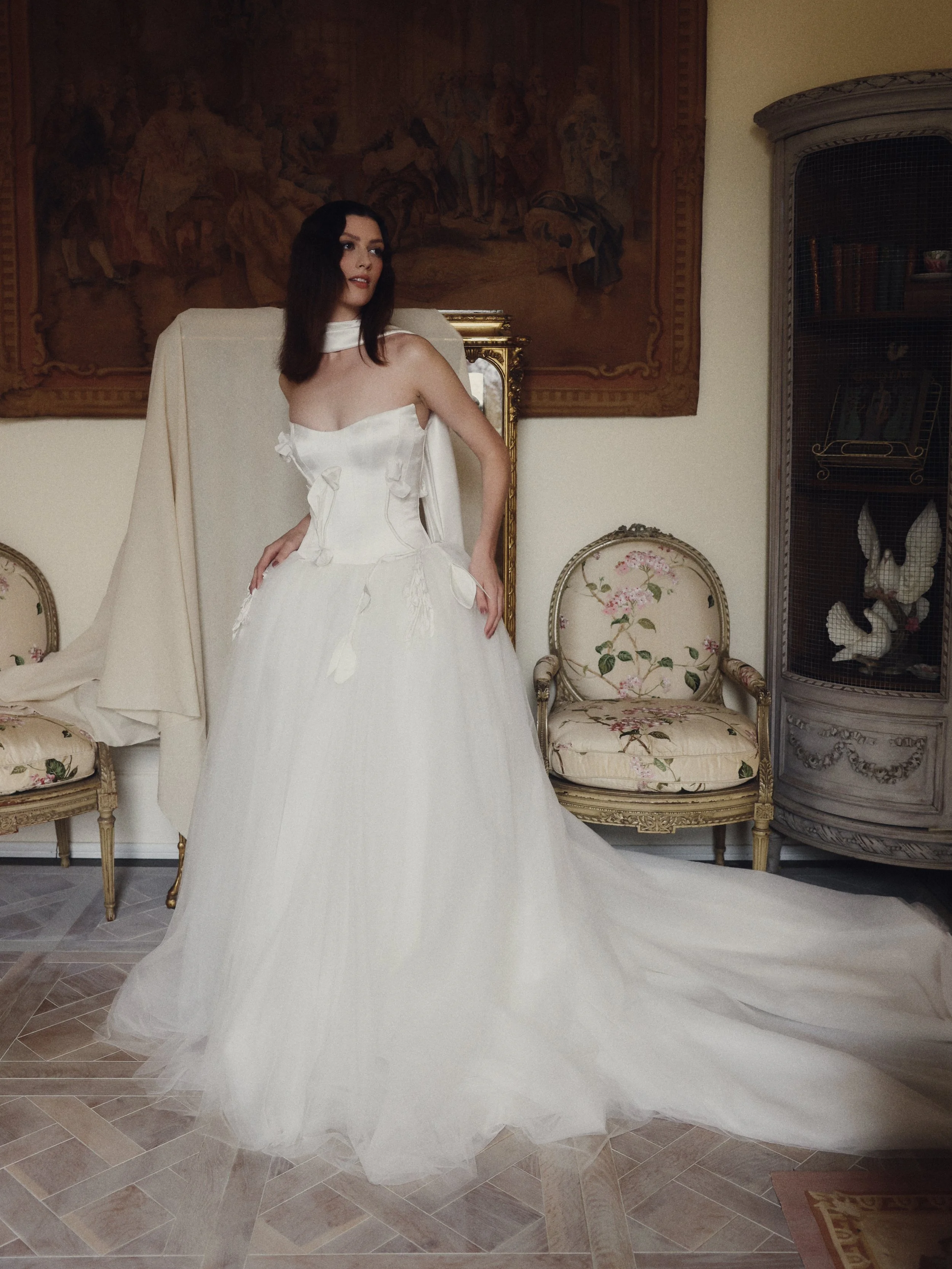 A woman in an elegant white wedding dress standing in a vintage-style room with floral chairs, a large tapestry, and antique furnishings.