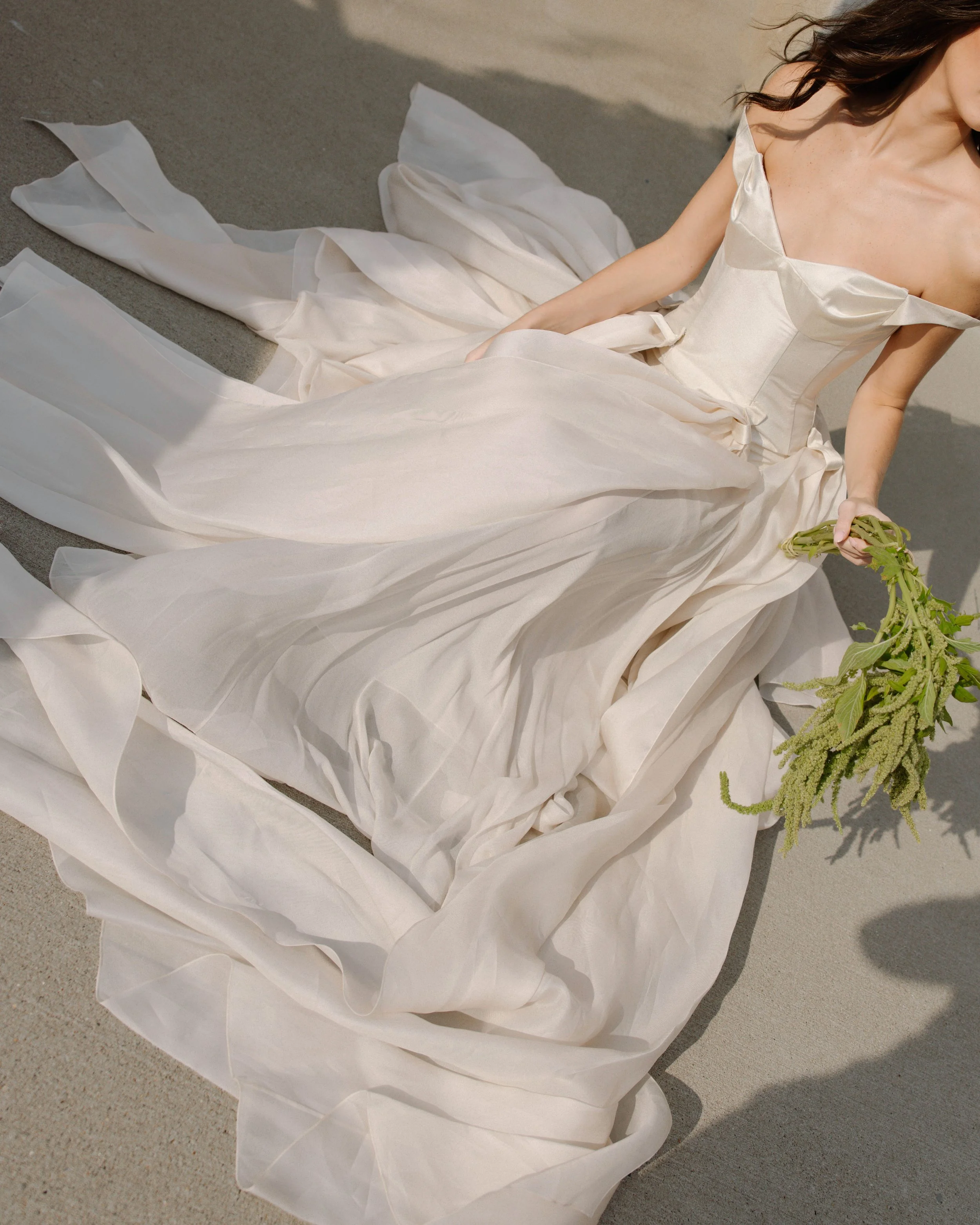 Woman wearing an off-shoulder white satin gown sitting on sandy ground, holding a green plant.