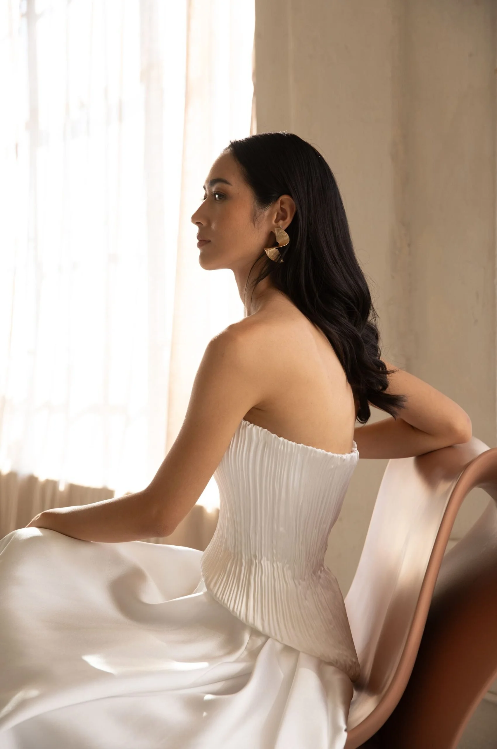 A woman with dark wavy hair sitting on a peach-colored chair near a window with sheer curtains, wearing a strapless white dress with pleated details and large gold earrings.