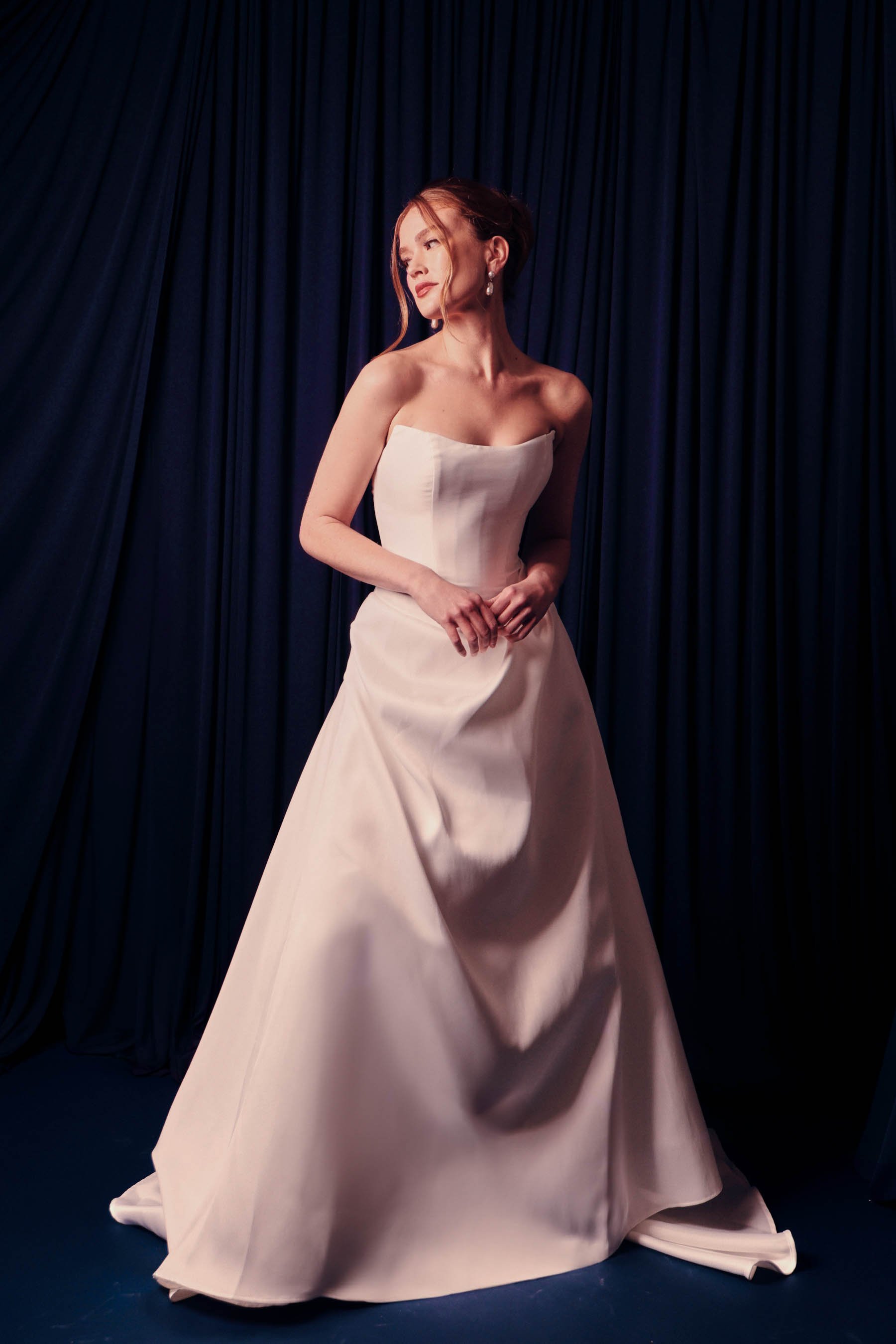 A woman in a strapless white gown standing in front of dark blue curtains.