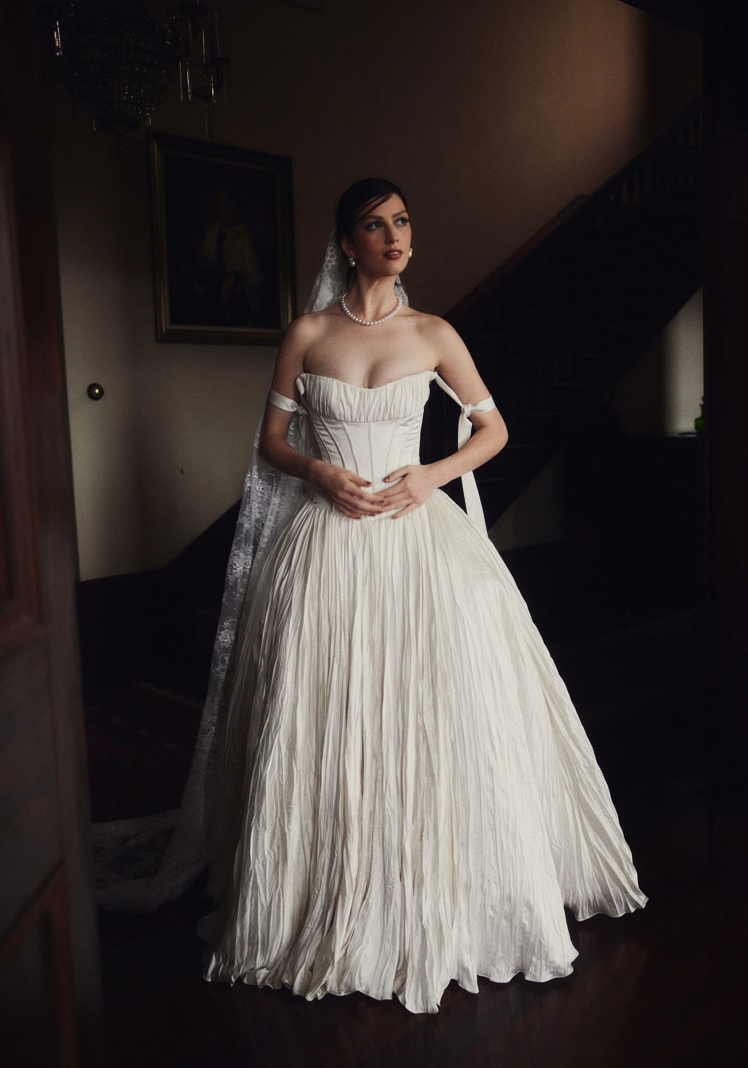 A woman in a vintage wedding dress with a pleated full skirt and strapless top, wearing a pearl necklace and earrings, with a lace veil, standing indoors near a staircase and a framed portrait on the wall.