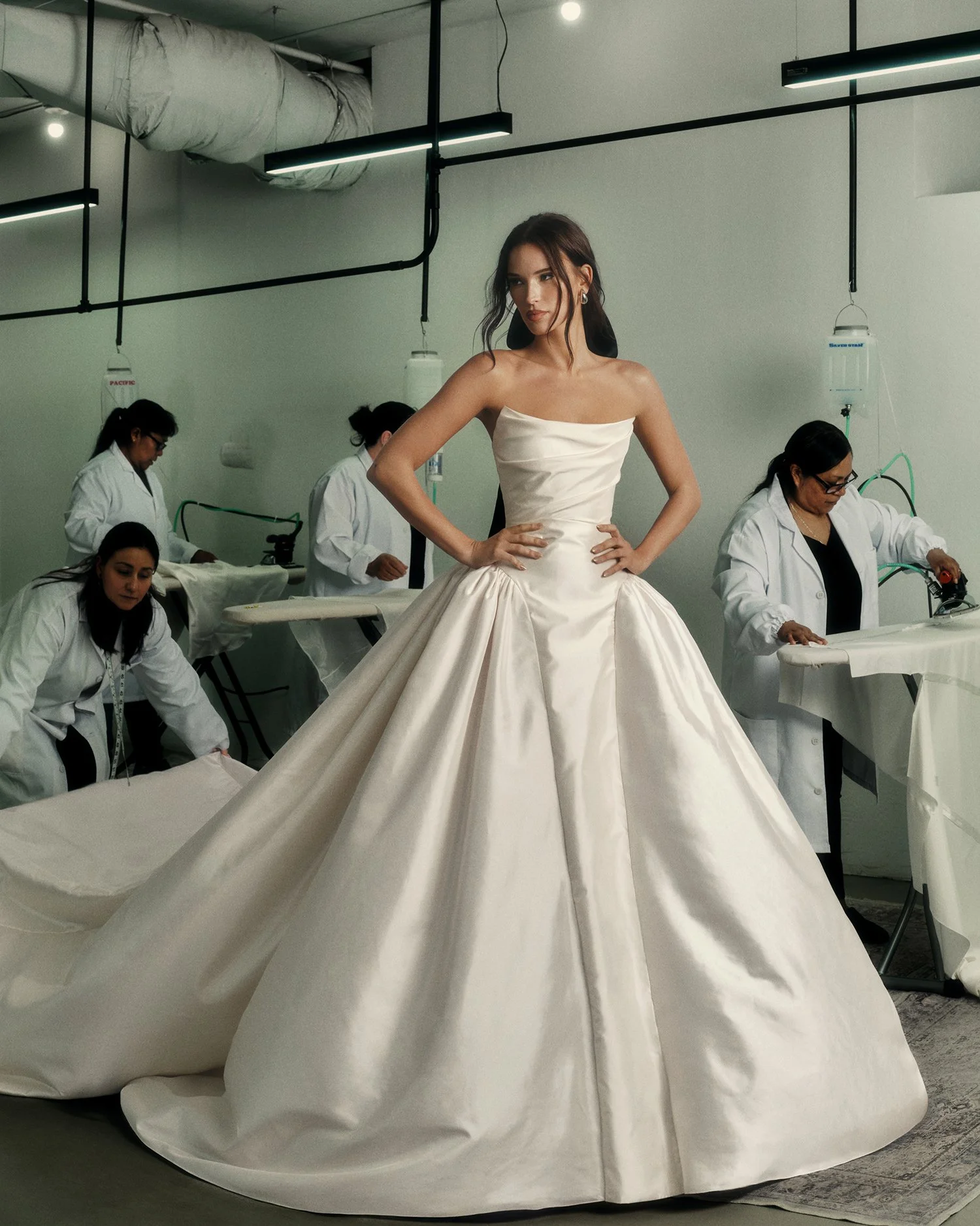 A woman in a white wedding dress standing with hands on her hips in a sewing or tailoring workshop, with several workers sewing or working on fabric in the background.