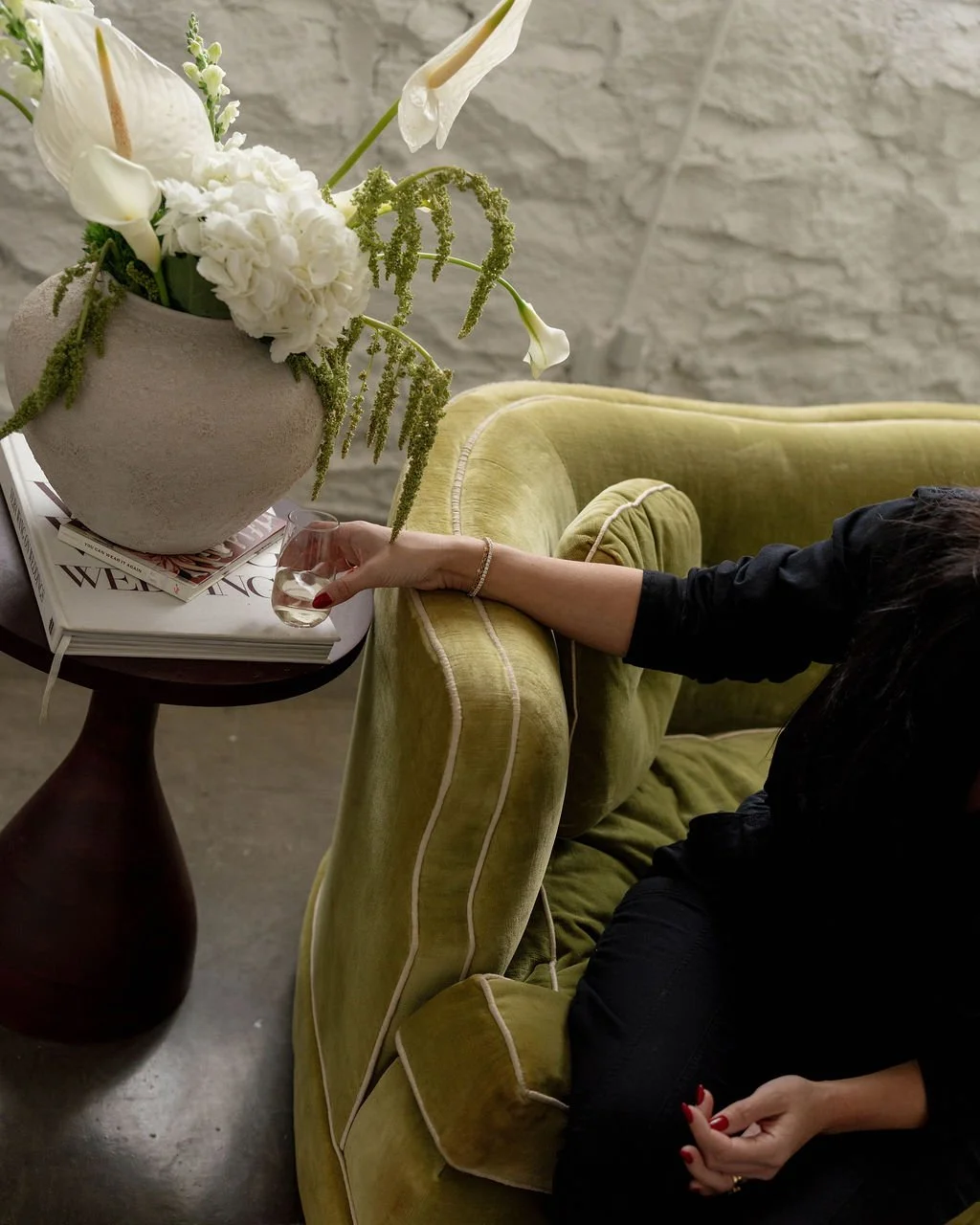 A woman's hand holding a glass of wine resting on a green velvet sofa, next to a side table with a large flower arrangement and stacked magazines, in a room with a brick wall background.