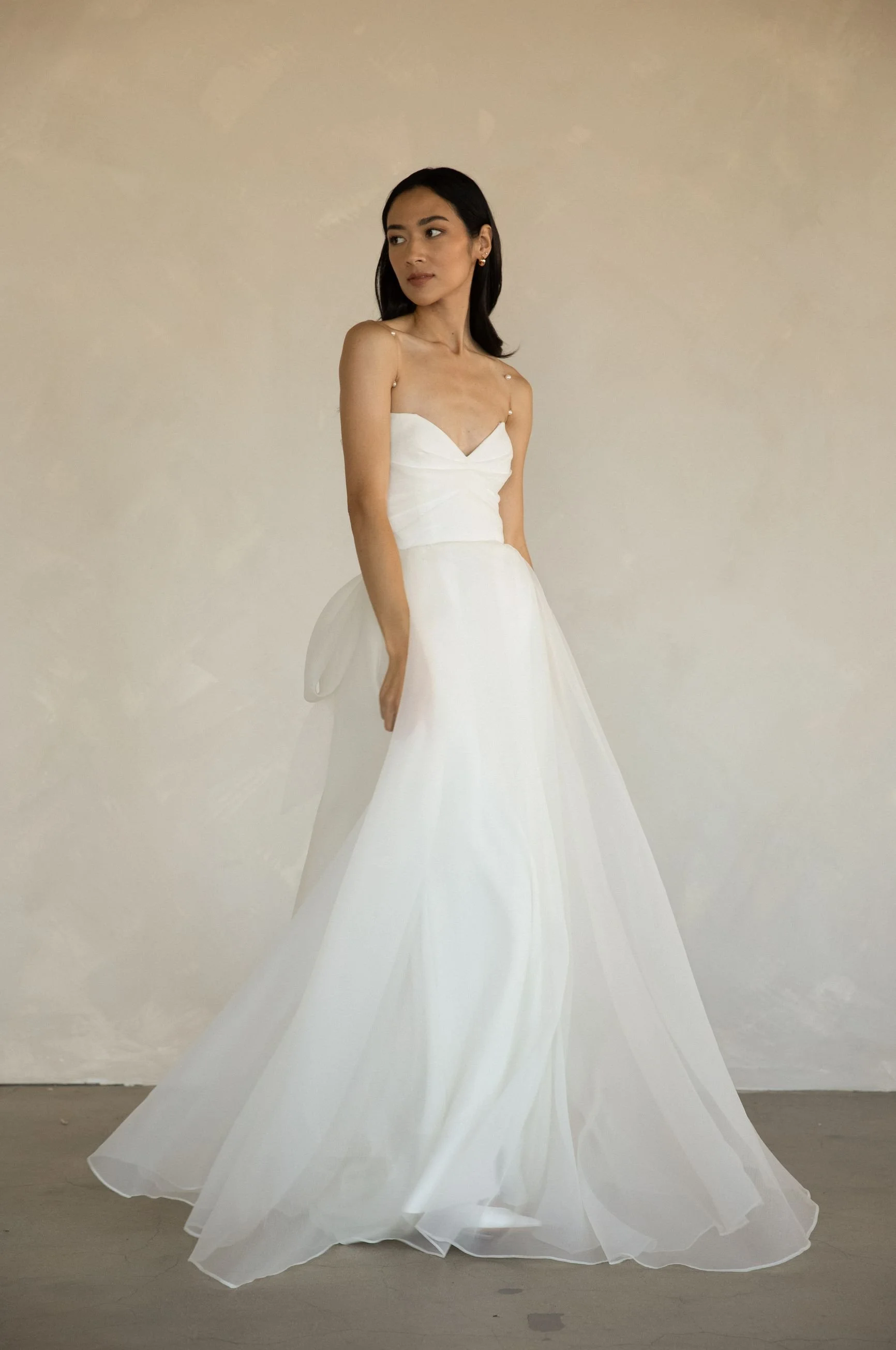 A woman in a white strapless gown with delicate shoulder straps standing against a plain beige wall.