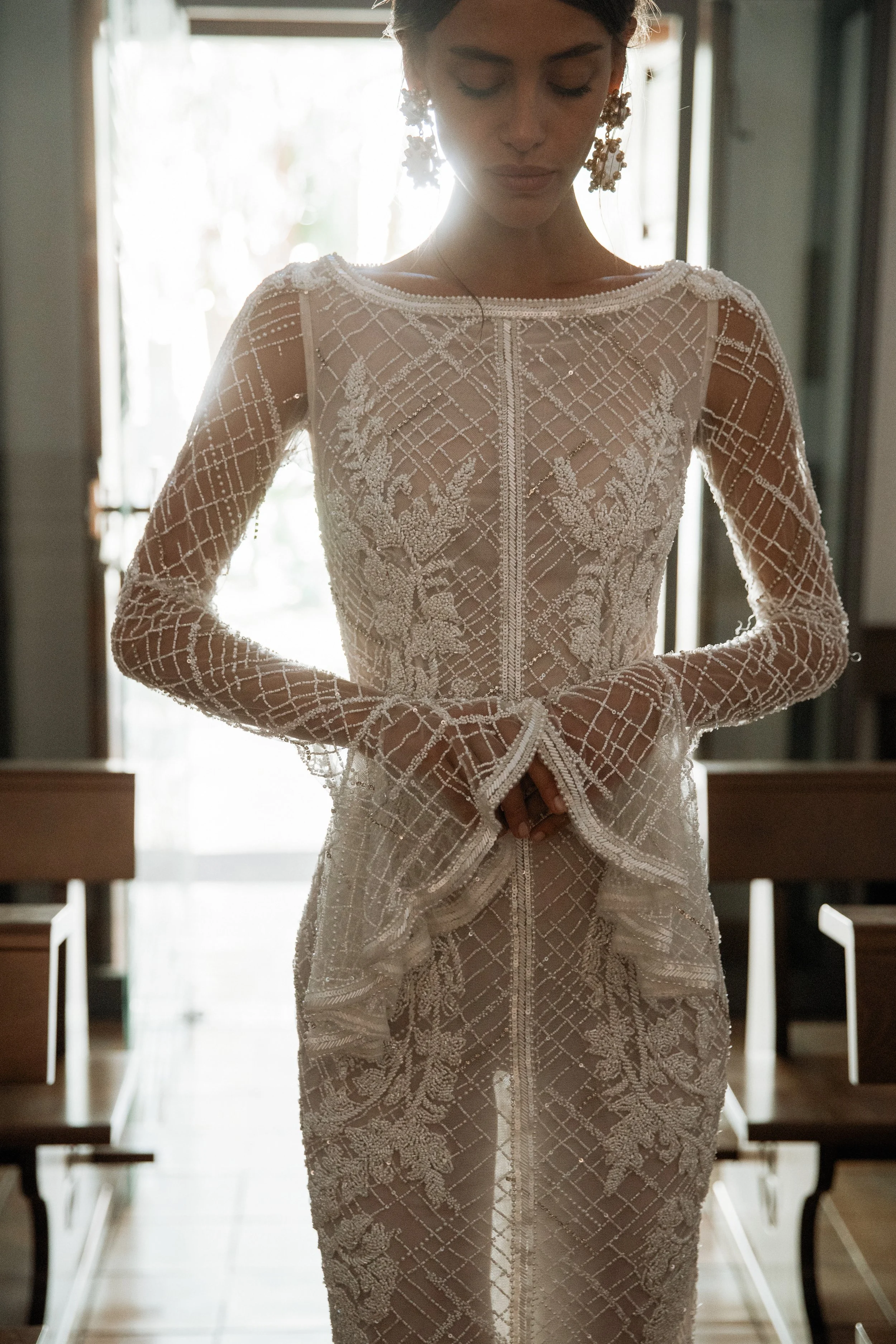 Woman in a detailed lace wedding dress standing indoors in front of a window.