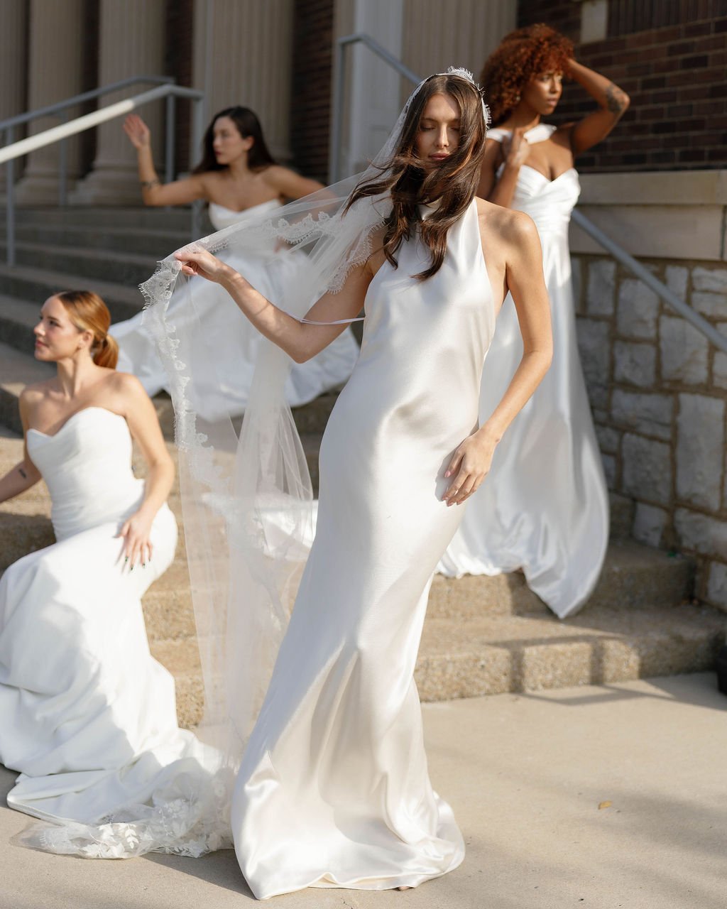 Four women in white bridal gowns standing on outdoor stairs, with one woman in a short dress sitting on the stairs, one woman standing with a veil, and another woman with curly hair adjusting her hair.
