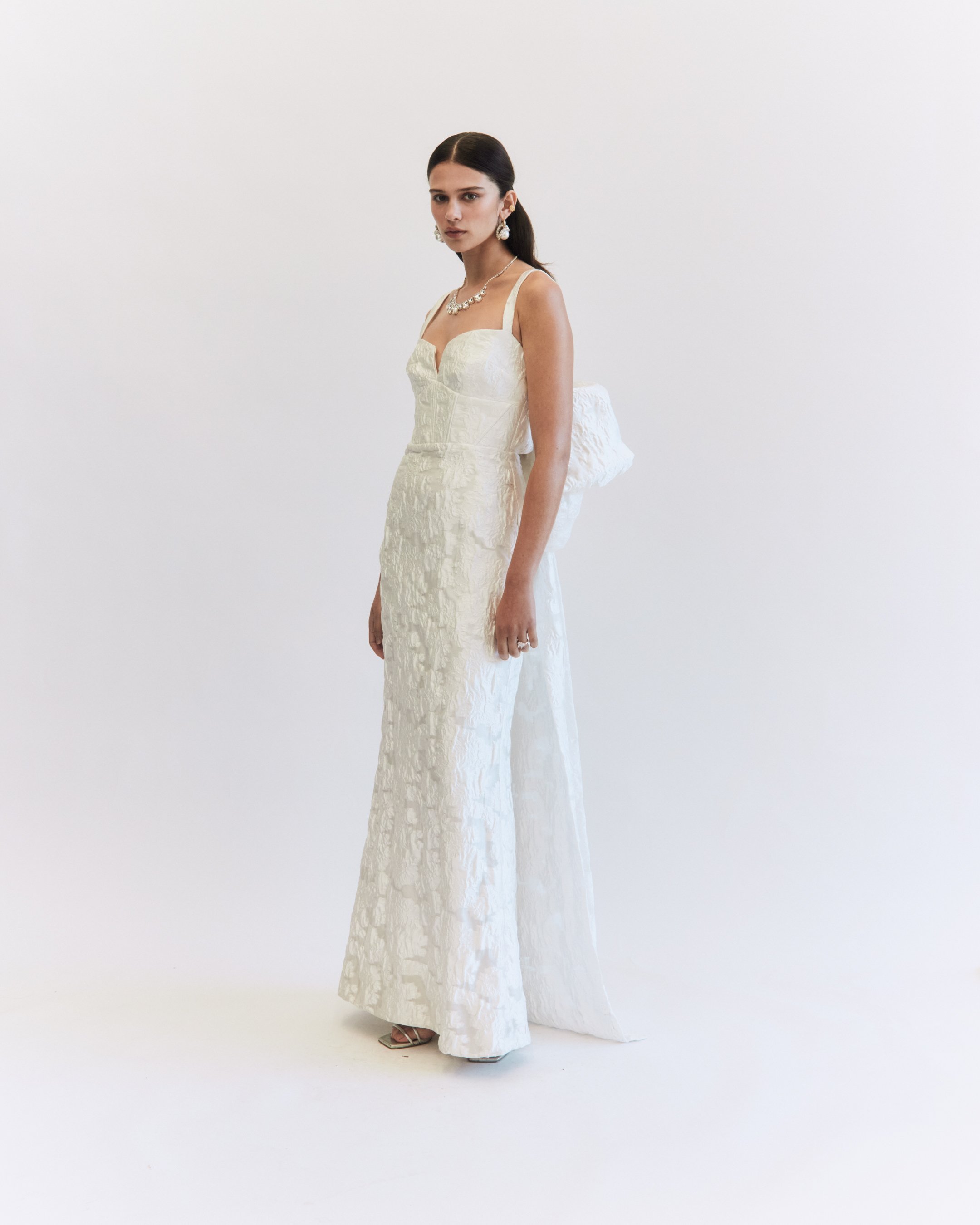 Young woman in a white lace gown with spaghetti straps, wearing pearl jewelry and holding a matching white clutch bag against a plain white background.