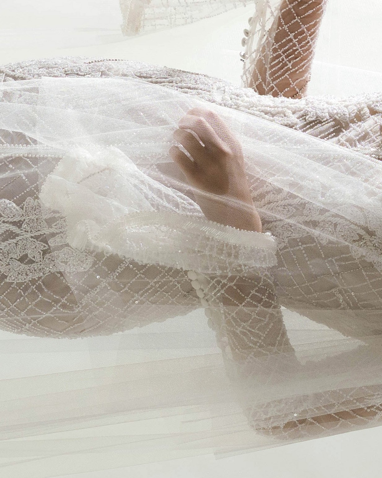 Close-up of a hand and a foot resting on a white, embroidered lace fabric, seen through sheer white curtain with sparkling details.