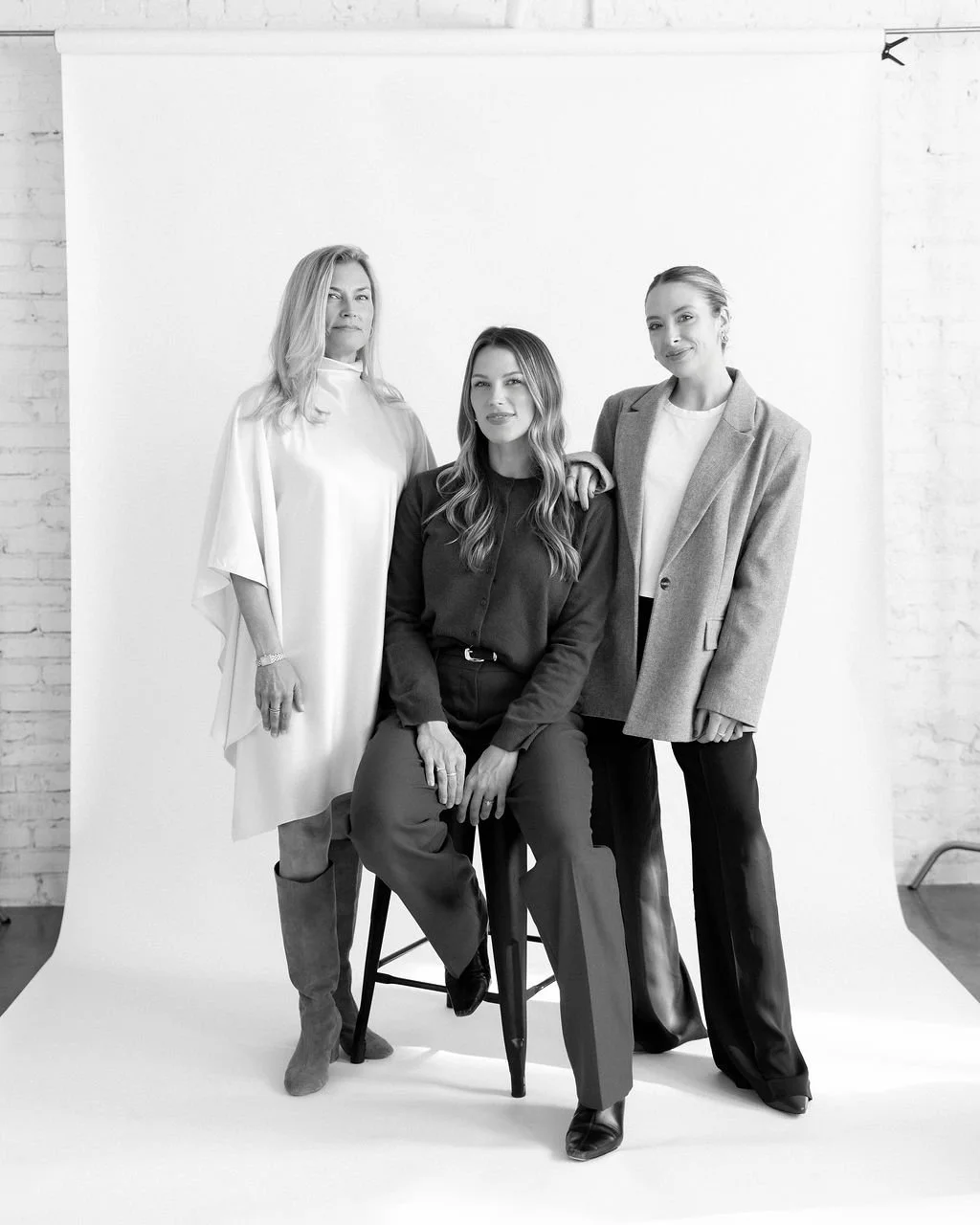 Three women are posing together in front of a white backdrop, with a brick wall visible on the sides. The woman on the left is wearing a light-colored dress and knee-high boots, the woman in the middle is seated on a stool wearing a dark blouse and trousers, and the woman on the right is wearing a blazer over a top and trousers. The image is in black and white.