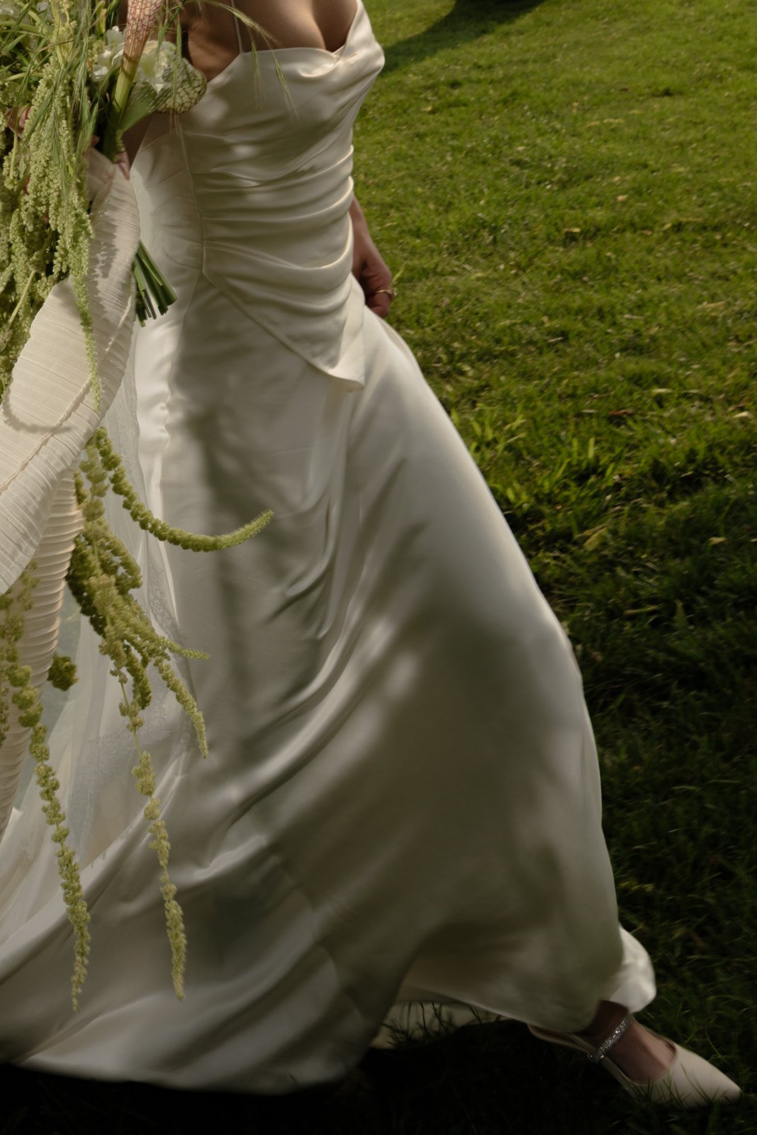 A person wearing a ivory satin wedding gown, holding a bouquet of greenery and flowers, standing on green grass in an outdoor setting.
