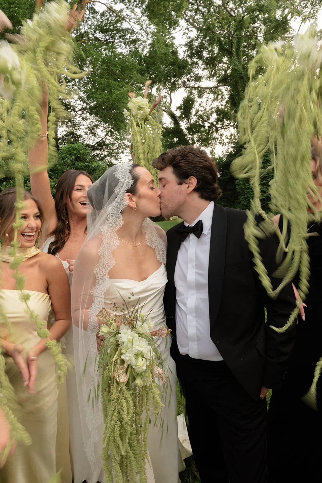 Bride and groom share a kiss surrounded by bridesmaids at an outdoor wedding, with greenery and hanging plants in the background.