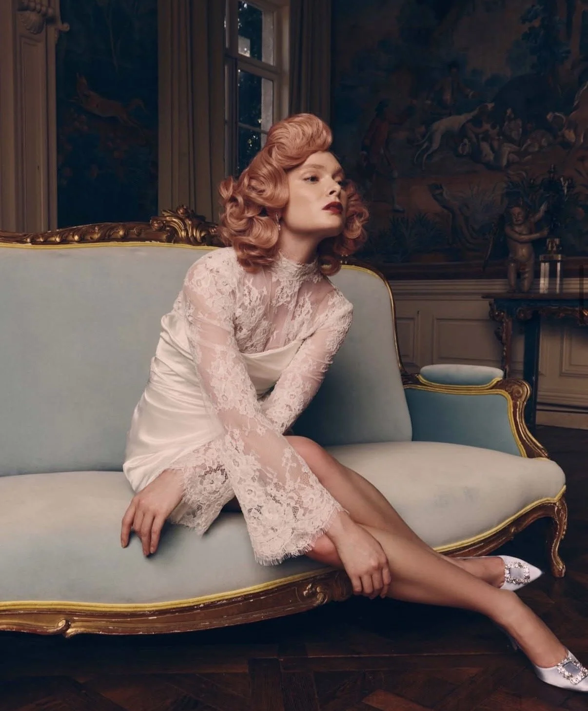 A woman with red, vintage-inspired hair and makeup, wearing a pale pink lace dress, sitting on a light blue antique sofa in an elegantly decorated room.