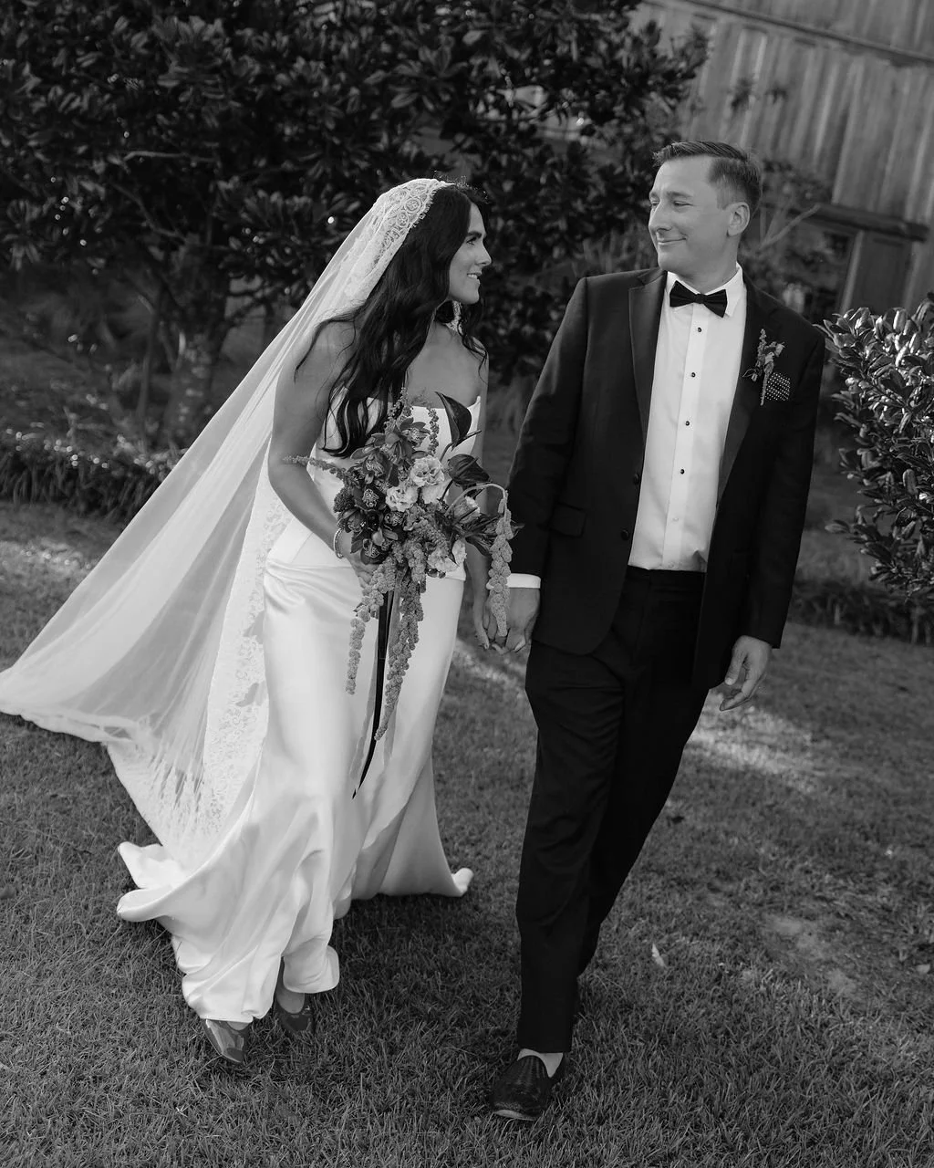 A bride and groom walking hand in hand on grass during their wedding, smiling at each other. The bride is wearing a wedding dress and veil, holding a bouquet. The groom is in a tuxedo with a bow tie. The background features trees and a wooden fence.