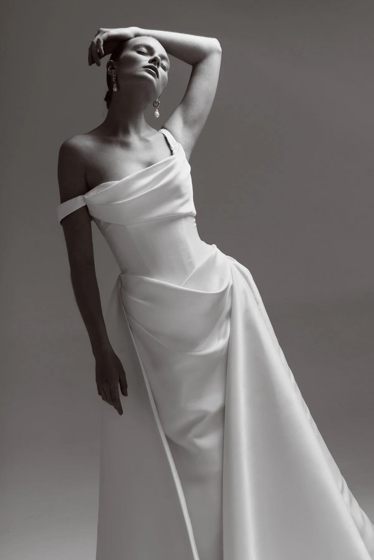 A woman in a strapless white satin gown with pleats, posing with one arm behind her head and the other by her side, against a plain gray background.