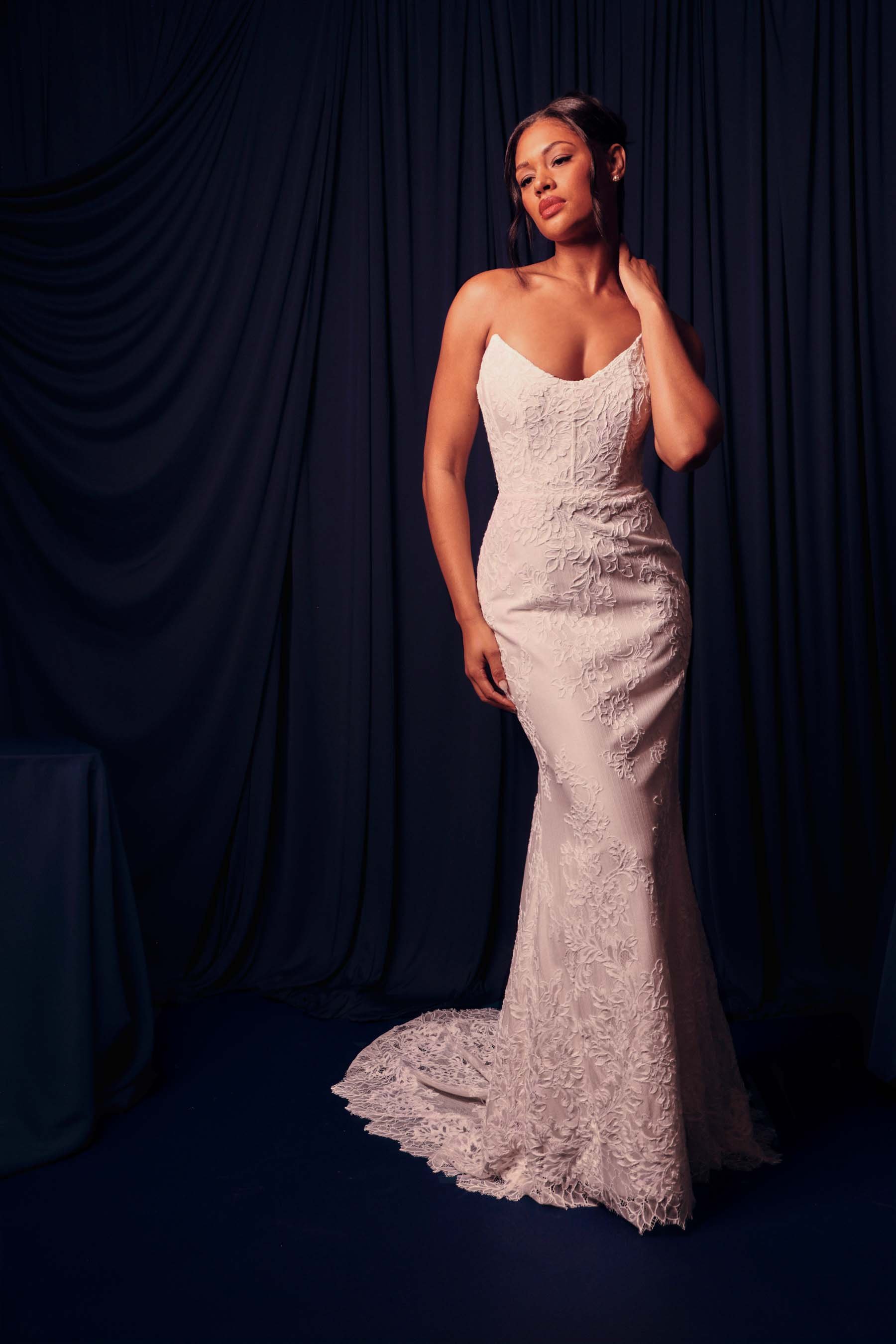 Woman in a white lace dress standing against a dark backdrop, looking contemplative.
