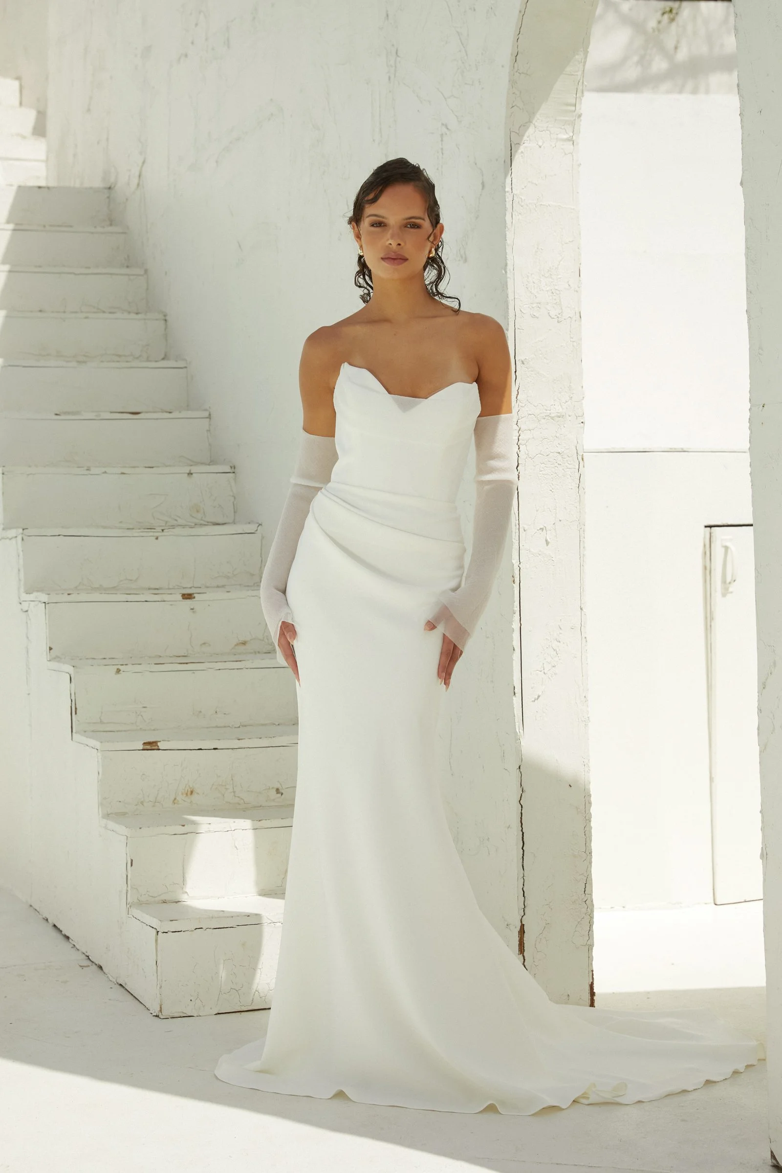 A woman in a strapless white wedding dress with detached sleeves posing against a white textured wall and stairs.
