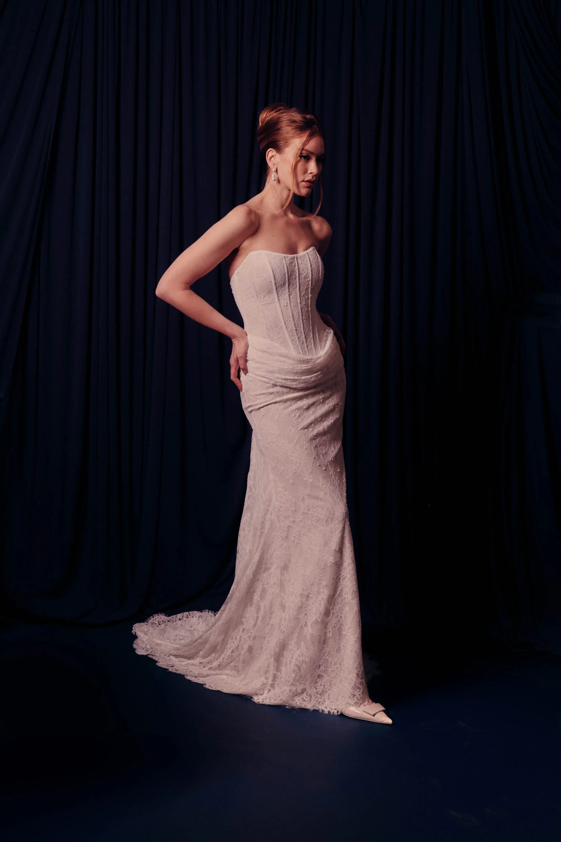 A woman in a strapless white lace wedding gown standing in front of dark curtains.