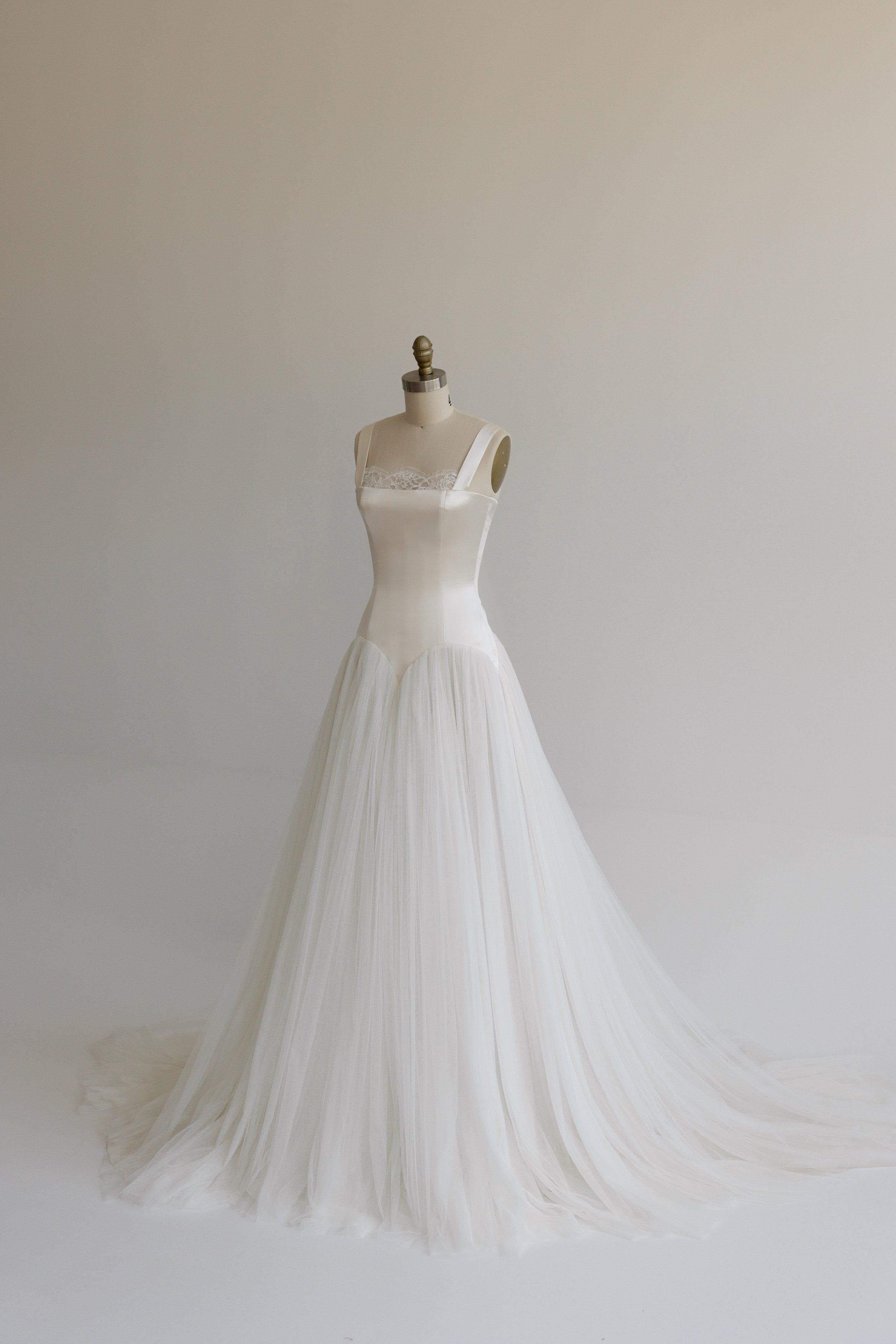 A white wedding dress on a mannequin with lace details on the bodice, a fitted waist, and a flowing, pleated skirt.