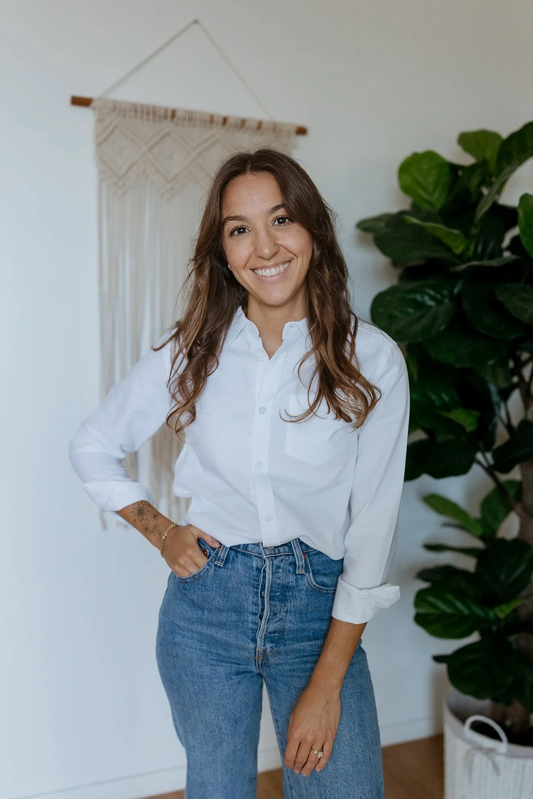A woman with long wavy brown hair smiling, wearing a white button-up shirt and blue jeans, standing indoors near a large green houseplant and a decorative wall hanging.