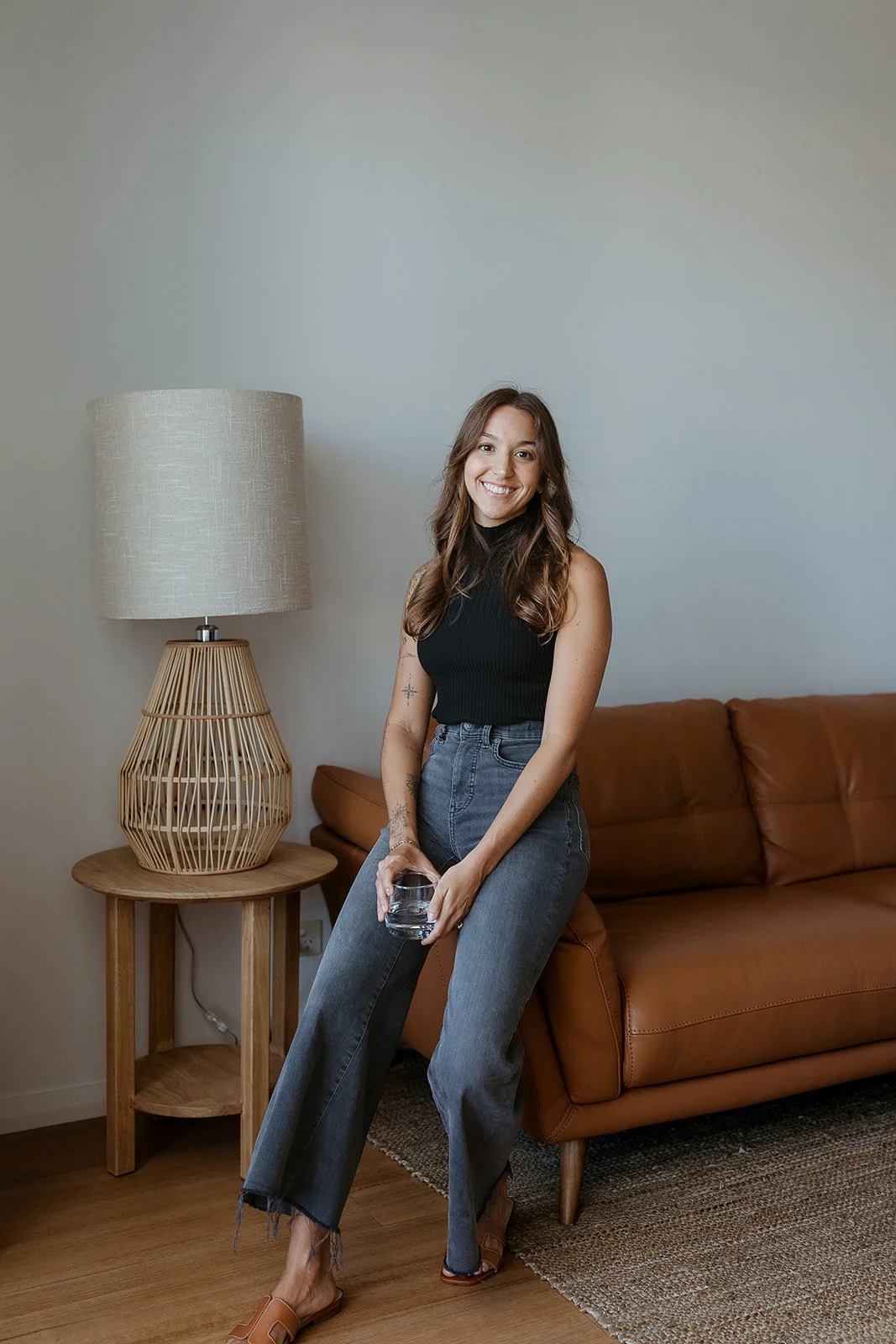 A woman with long brown hair smiling, wearing a black sleeveless top and high-waisted jeans, holding a glass of water, standing next to a brown leather couch and a wooden side table with a beige lamp, in a cozy living room.