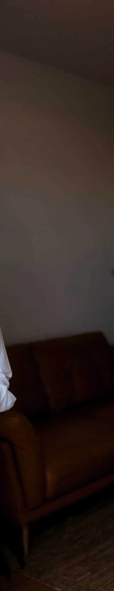 Part of a brown sofa and a white shirt visible in a dimly lit room with plain walls.