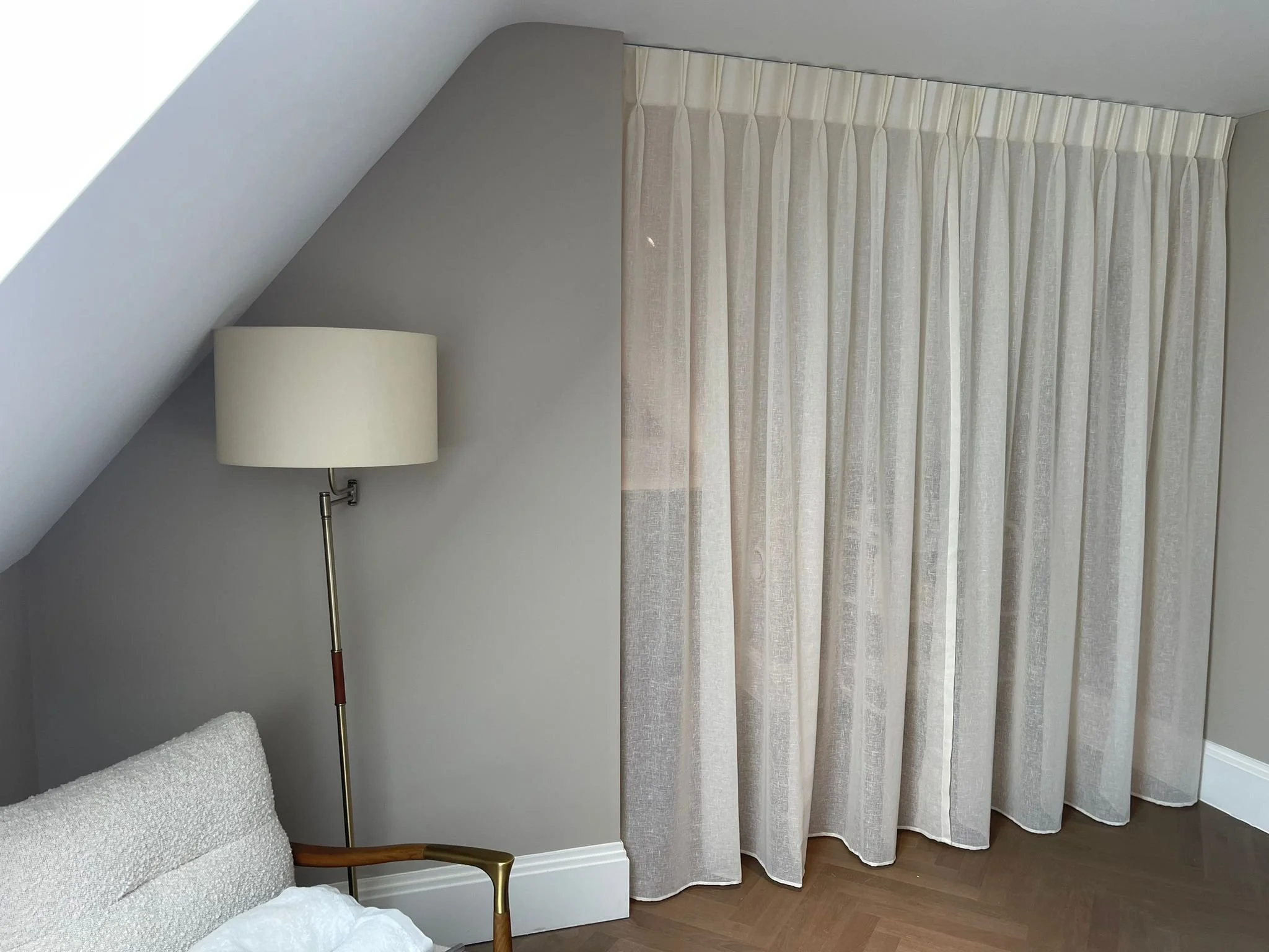 View of a corner in a bedroom with a sloped ceiling, a tall floor lamp with a beige shade, part of a white textured bed, and beige curtains covering a window or closet.