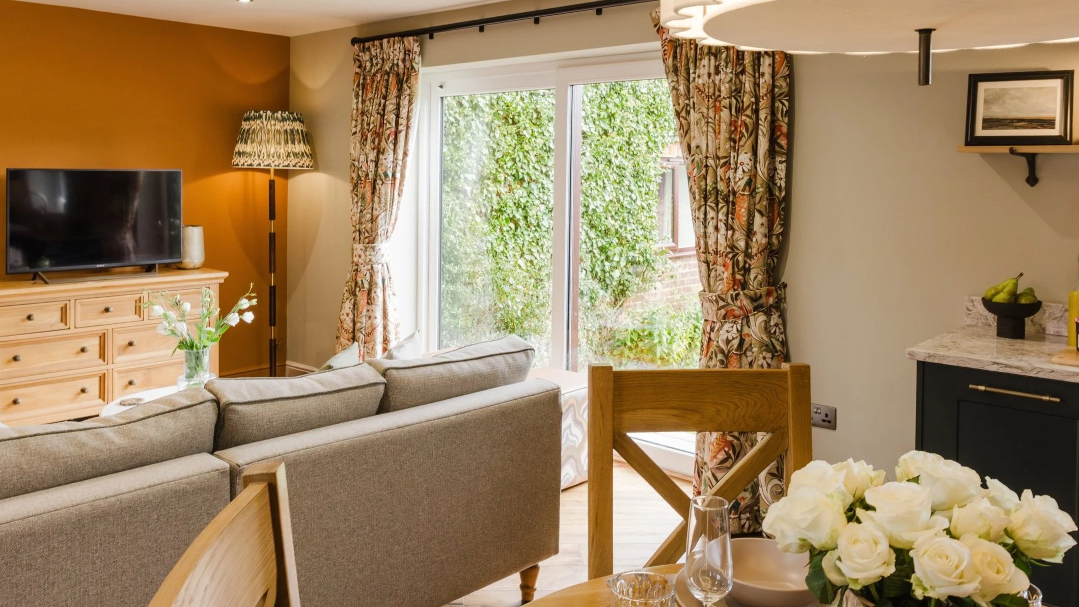 Living room with beige sofa facing a wooden dresser with a television, standing lamp, and a vase of white flowers. Large sliding glass door with floral curtains opens to a garden. Part of dining table with a floral centerpiece is visible.