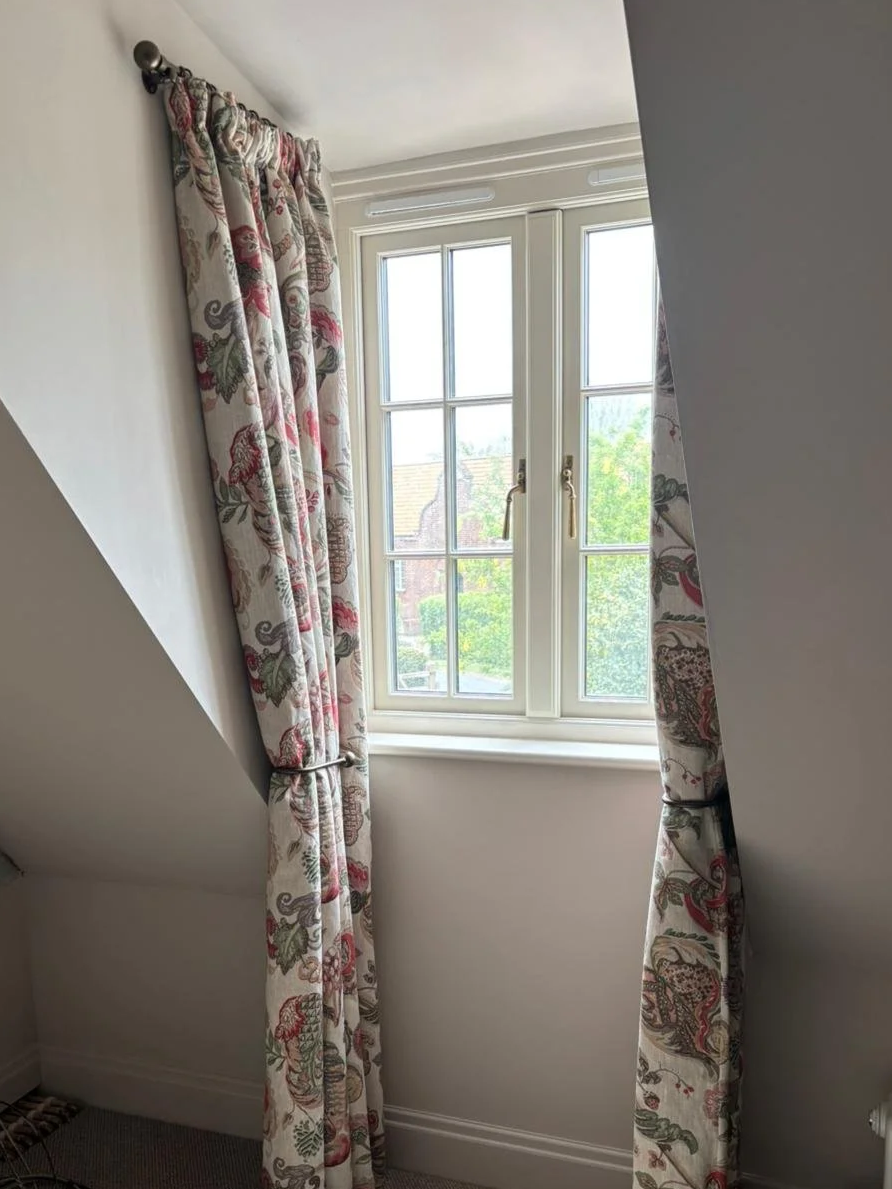 A window with cream-colored frames and a latch, partially open, with floral patterned curtains in shades of pink, green, and beige on a rod, letting in natural light.