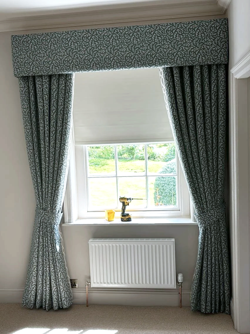 A window with patterned curtains and a ceiling valance in a room, with a cordless drill, a yellow box, and a small container on the windowsill, and a radiator below the window.