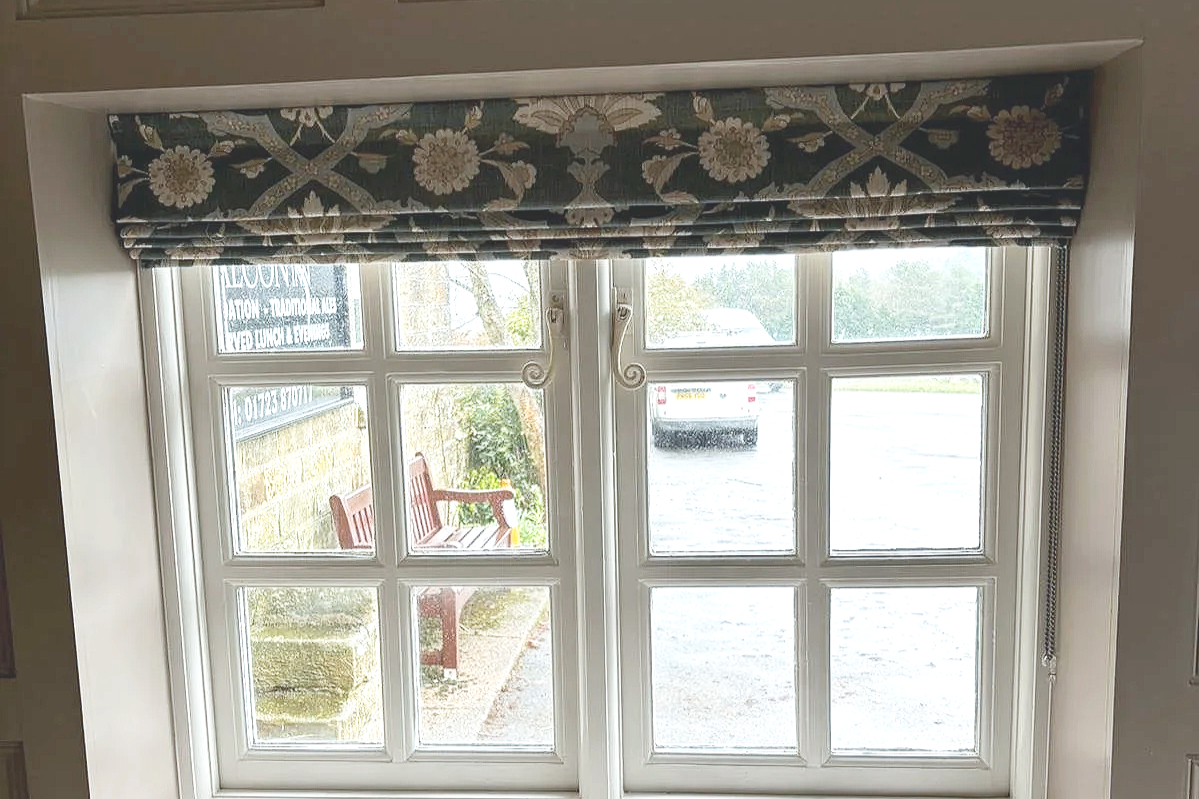 View of a window with a blue floral curtain valance, outside a brick wall and a wooden bench, with a car parked in the distance.