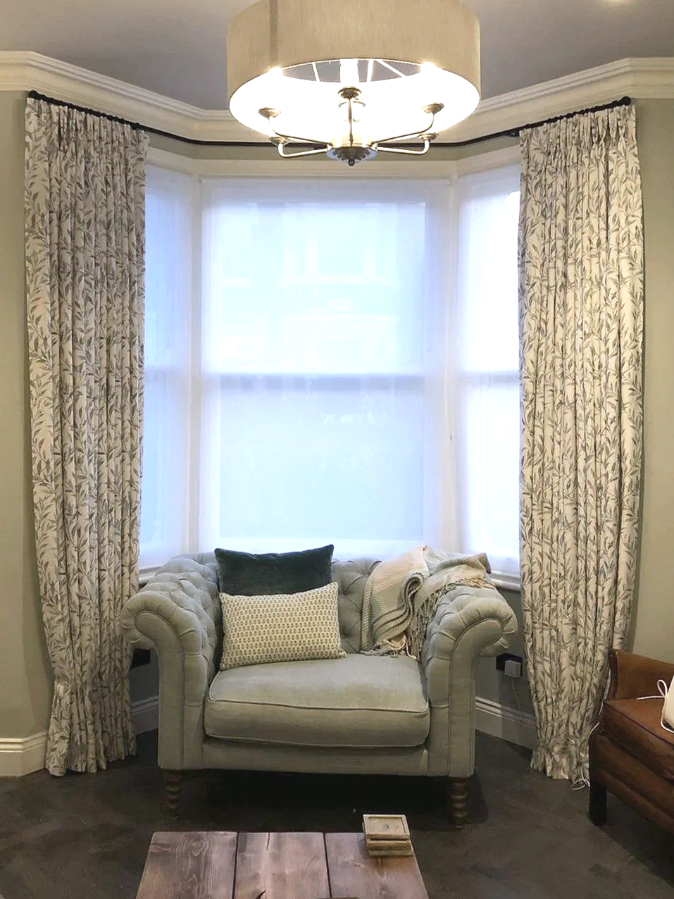 A cozy reading nook with a plush beige tufted loveseat decorated with two pillows, one navy and one patterned, topped with a blanket. The nook is situated in a bay window with sheer white curtains and patterned drapes on each side. A wooden coffee ta