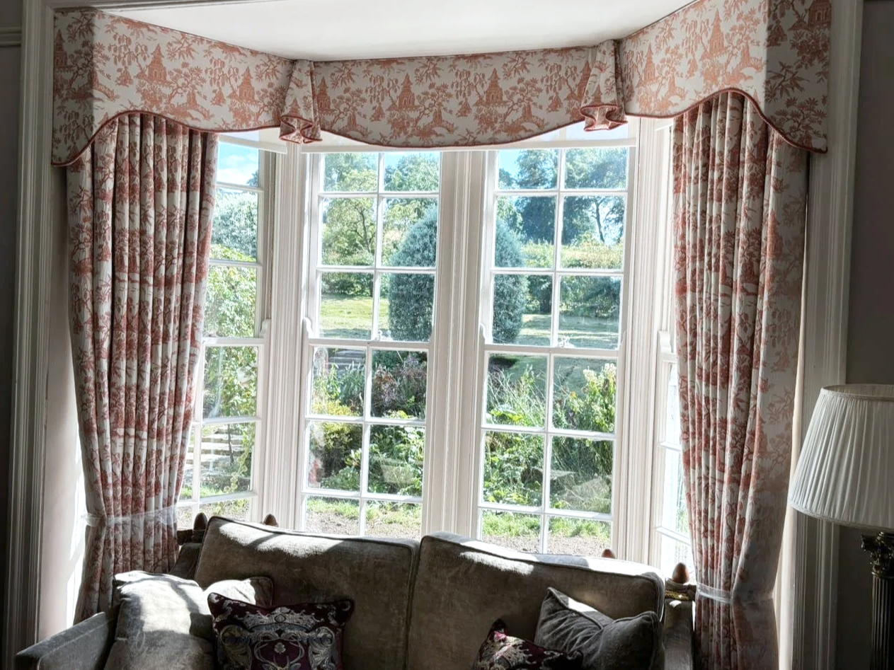 A living room with large bay windows dressed with floral pink and white curtains, overlooking a garden with trees and bushes.
