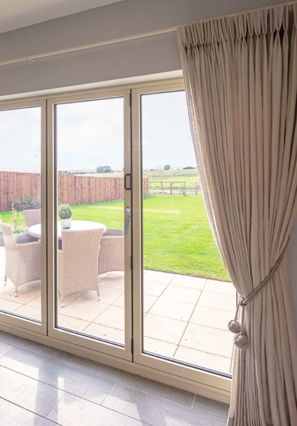 View of a backyard outdoor patio with wicker chairs and a table, seen through large glass sliding doors with beige curtains, overlooking a grassy lawn and a wooden fence.