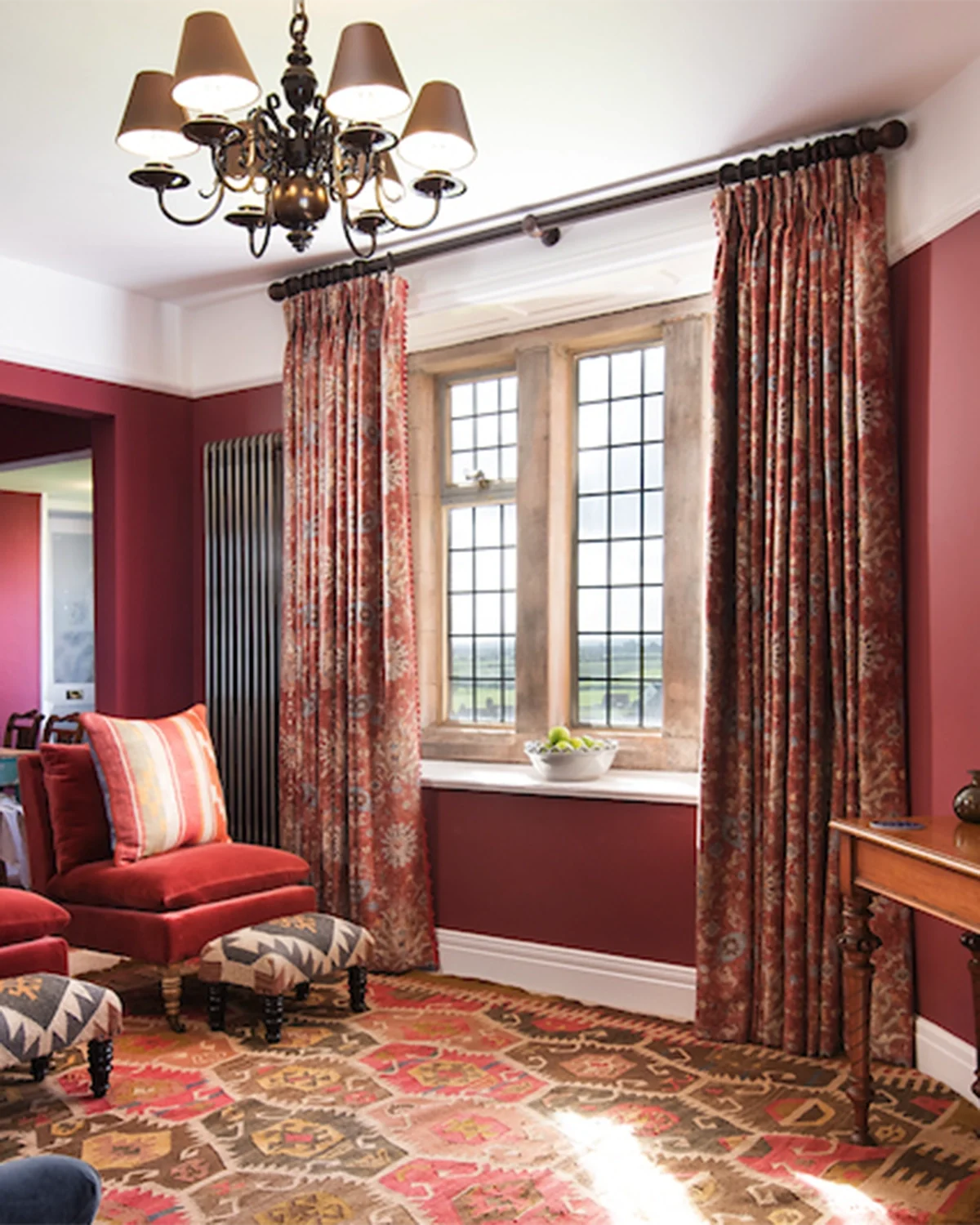 Living room with red walls, patterned curtains, a large window, and vintage furniture including red chairs and an ottoman, a chandelier, and a colorful rug.