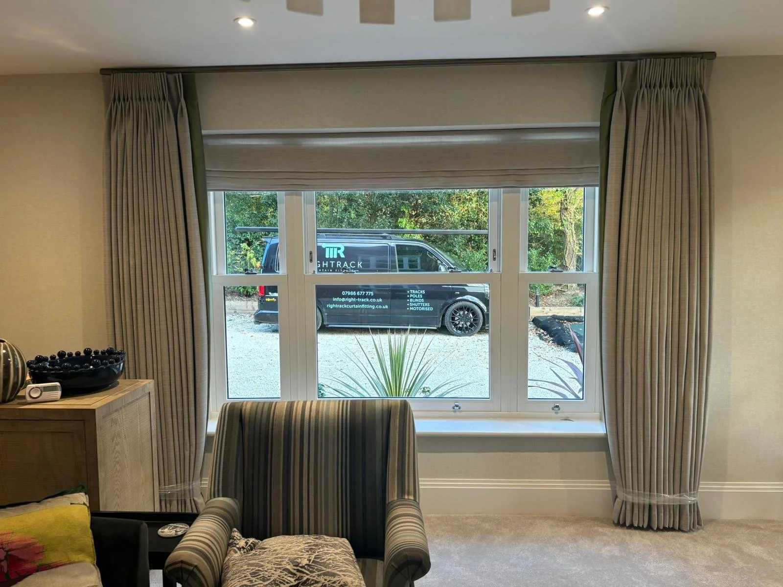 View through a house window showing a parked black van with white text, trees, and greenery outside. The window has beige curtains pulled back and a windowsill with decorative items inside.