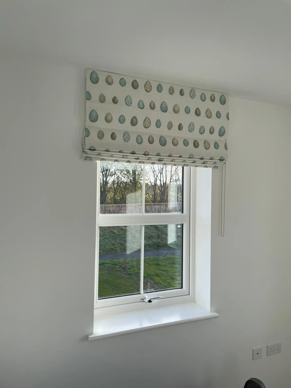 A room with a window featuring a decorative Roman shade with a pattern of colored eggs, overlooking a lawn with trees in the background.