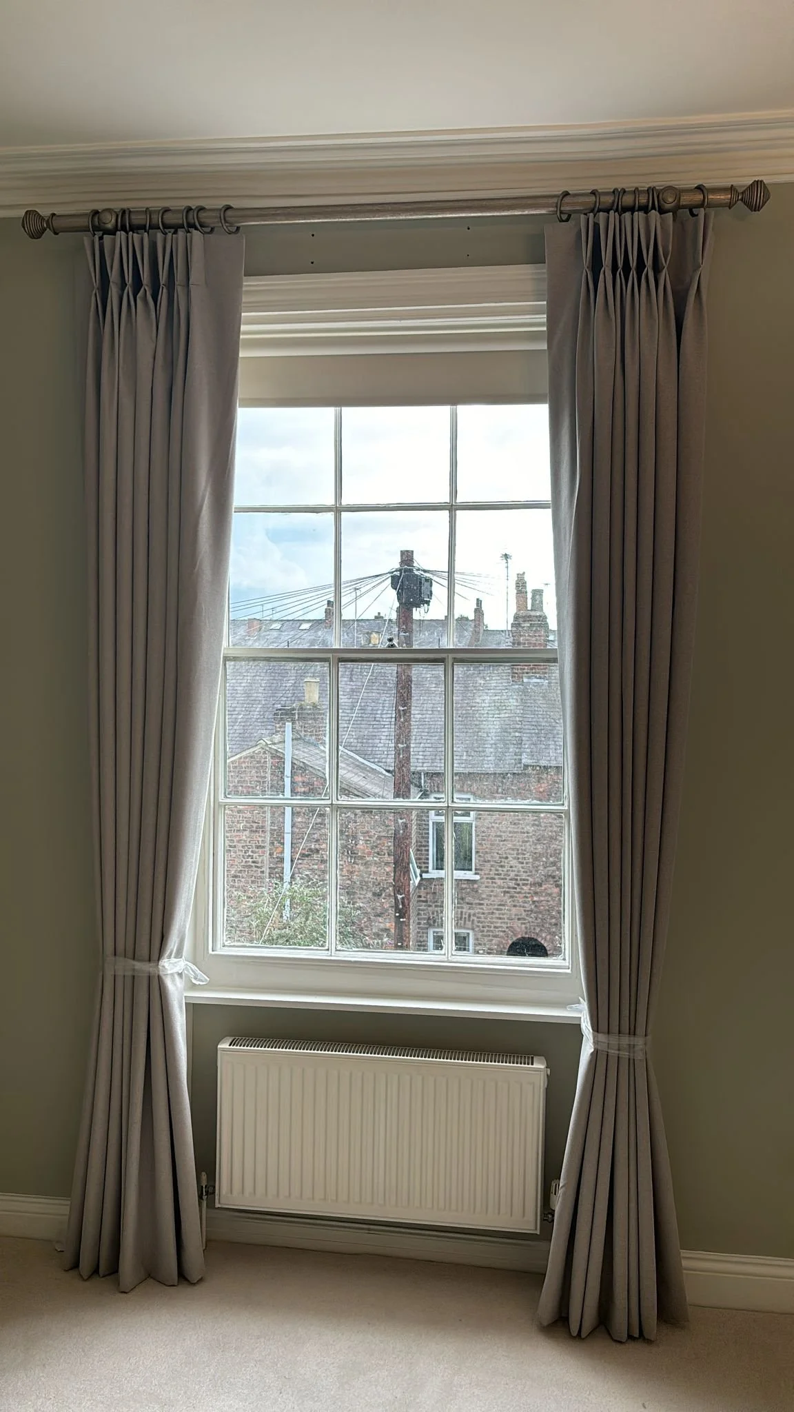 A window with beige curtains pulled to the sides, showing an outdoor view of a brick building, rooftops, a utility pole, and cloudy sky.
