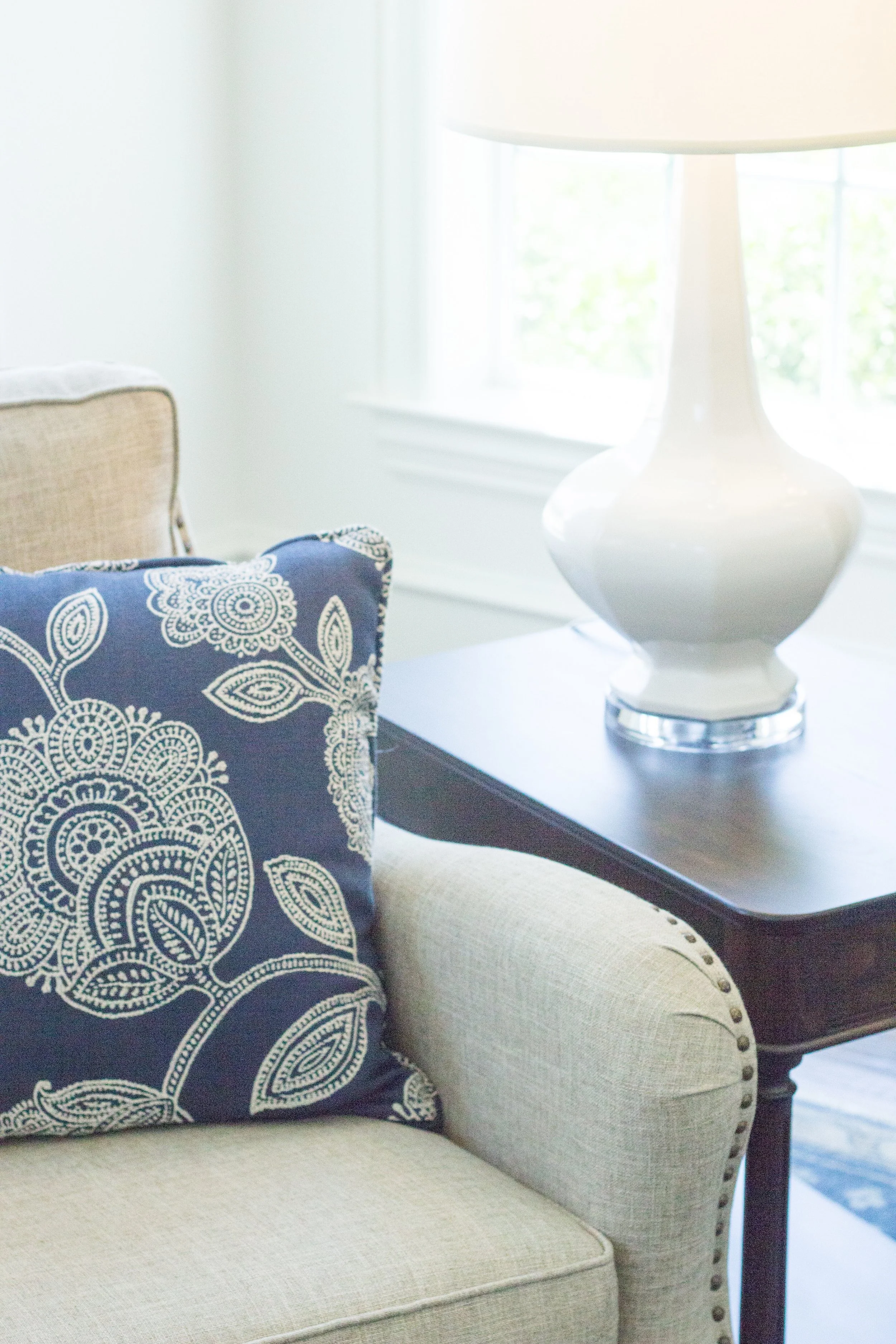Close-up of a beige armchair with studded trim, a dark wood side table, a white ceramic lamp, and a patterned blue and white pillow, near a window with bright natural light.
