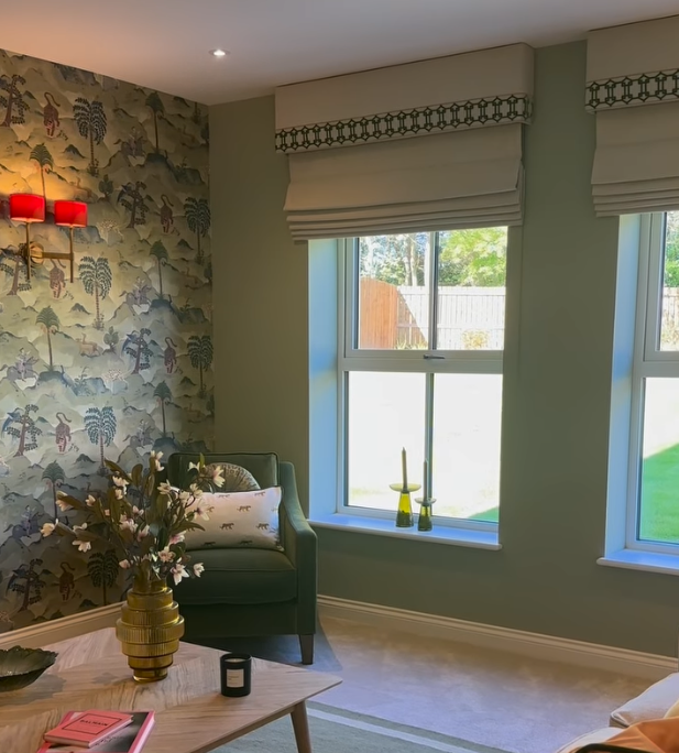 A cozy corner of a living room featuring a green armchair, floral wallpaper with trees and animals, large windows with white blinds, and a wooden table with a vase of flowers, candles, and books.