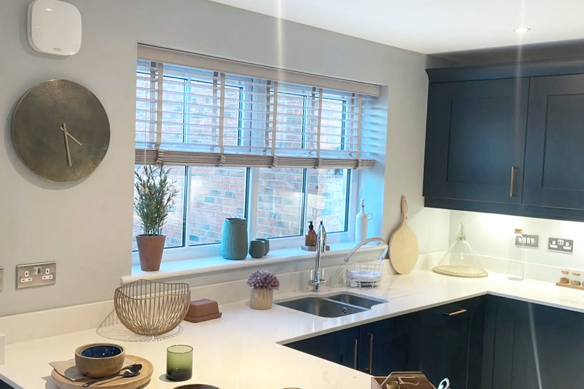 A modern kitchen with navy blue cabinets, a white countertop, and a window with blinds. There is a potted plant, decorative vases, a cutting board, and various kitchen items on the counter. A wall clock is on the left wall.
