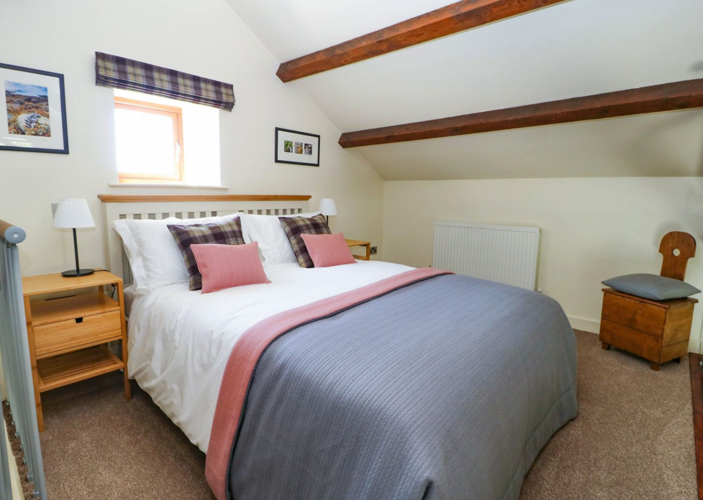 Cozy bedroom with a white bed, plaid pillows, pink pillows, and a pink and gray blanket. Wooden nightstands with white lamps, small window with plaid valance, art on the walls, and wooden ceiling beams.