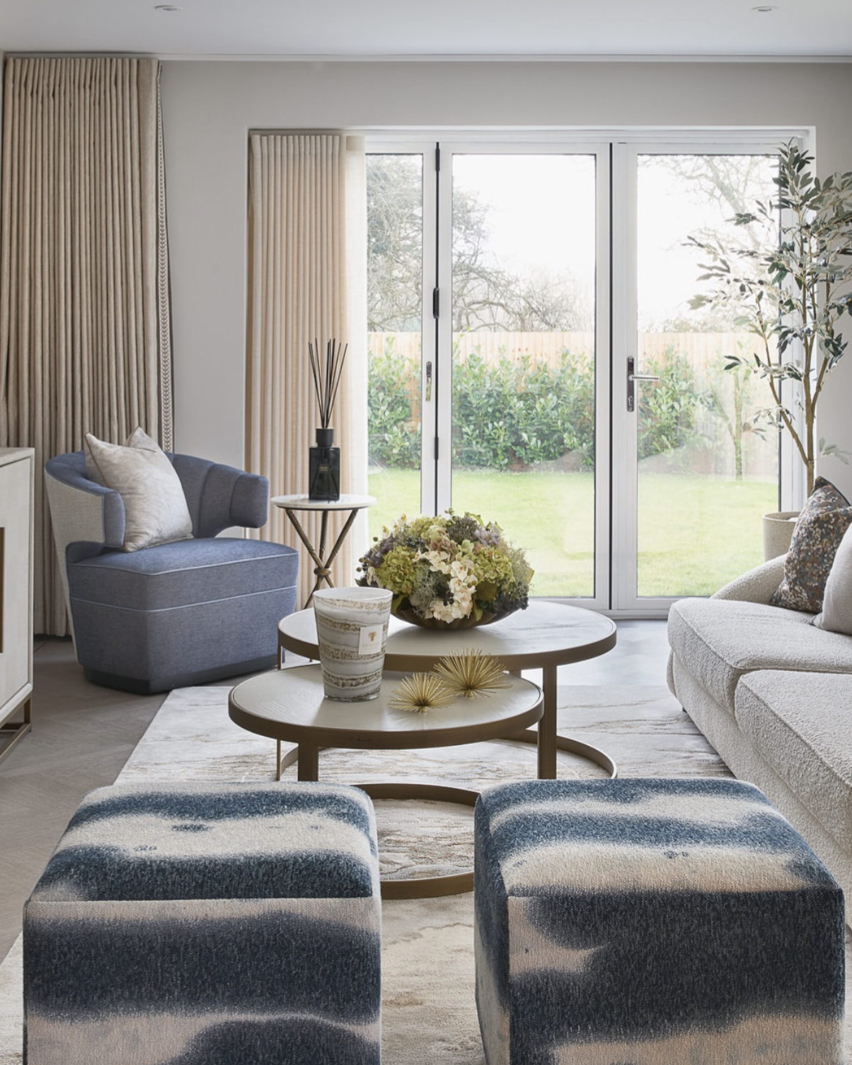 Living room with a beige sofa, gray armchair, two round coffee tables, two patterned ottomans, large sliding glass door, and outdoor garden view.
