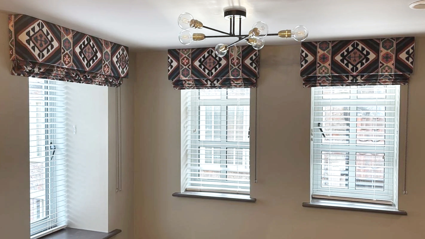 Three windows with white blinds and patterned valances in a room with cream-colored walls, illuminated by a modern chandelier with exposed bulbs.