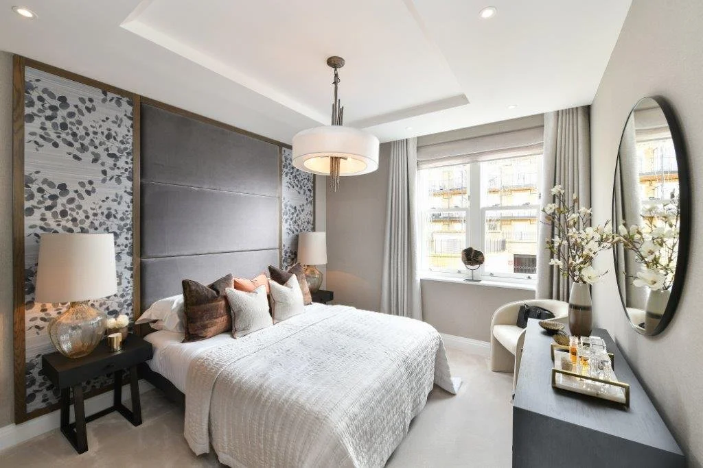 Modern bedroom with large bed, decorative pillows, nightstands with lamps, a chair near the window, and a dresser with decor and flowers, bright natural light from window.