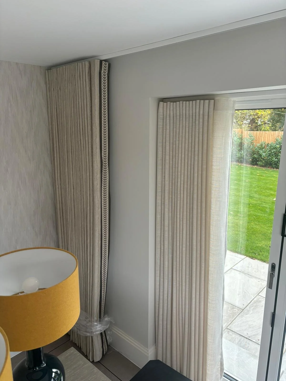 Interior of a room with a sliding glass door opening to a garden, with beige curtains and a table lamp with a yellow lampshade.