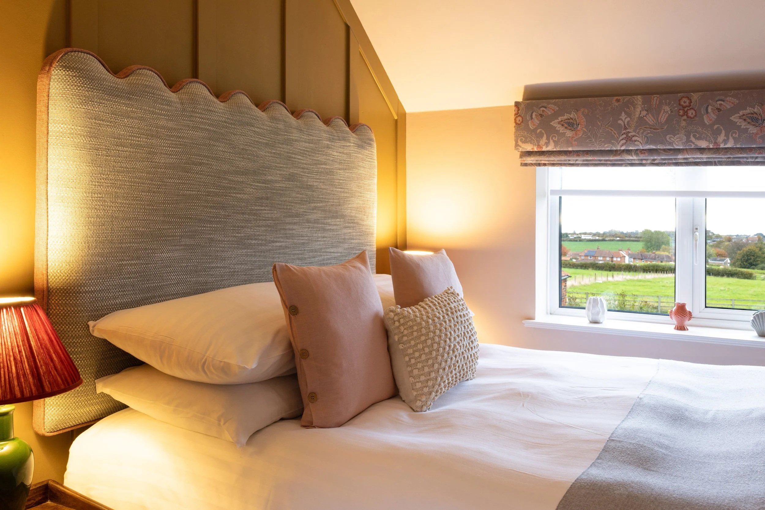 A cozy bedroom with a large bed, decorative pillows, a large textured headboard, a nightstand with a red lamp, and a window with a scenic view of green fields and houses.