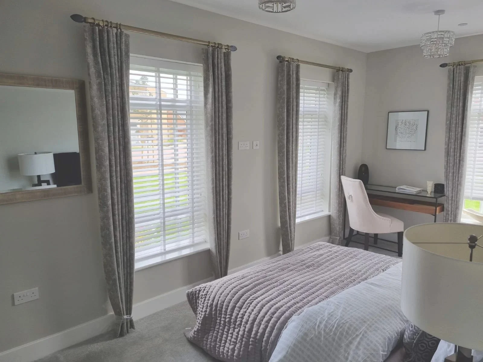 A bedroom with two large windows with white blinds and patterned gray curtains. A bed with a textured beige blanket and pillows is visible. A mirror on the wall reflects a lamp and chair. A small desk with a pink chair, some books, and decorative ite