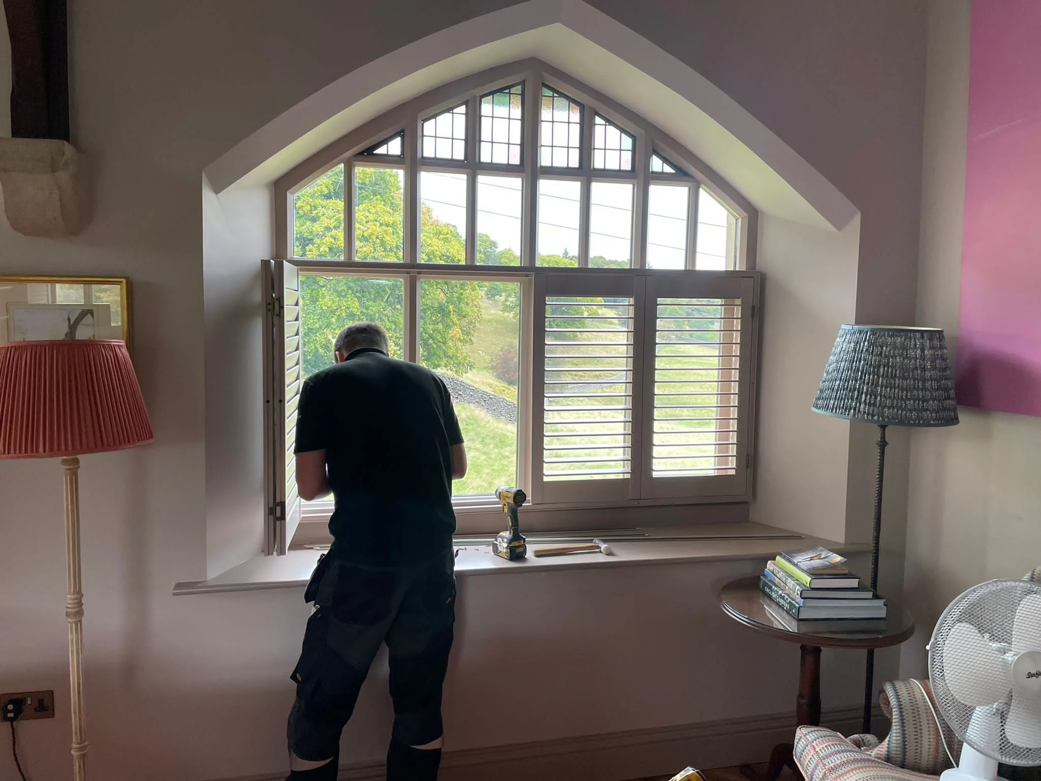 A person installing or repairing a large, multi-pane window in a room with sunlight coming through. The room has decorative lamps, a small round table with books, and striped armchair.